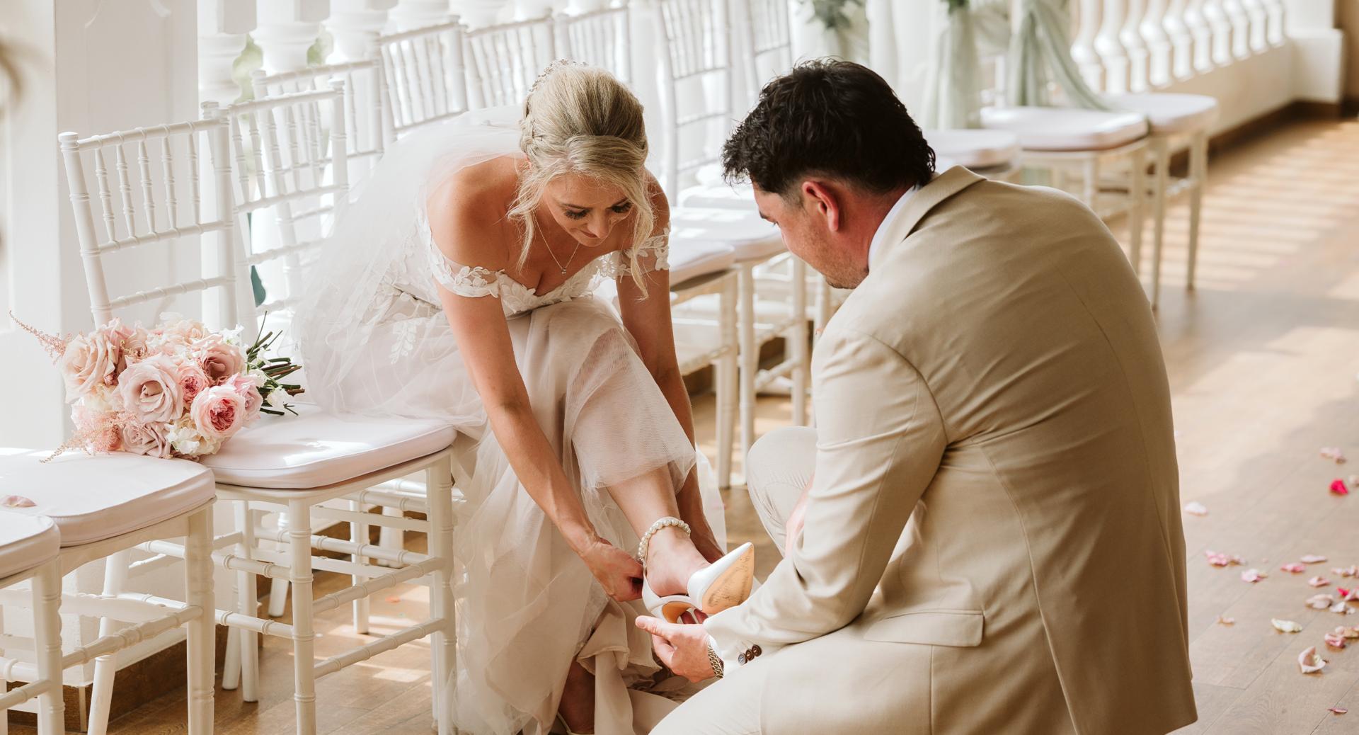 A bride sits on a white chair as a groom kneels to help her put on a shoe; a bouquet and scattered flower petals are nearby.