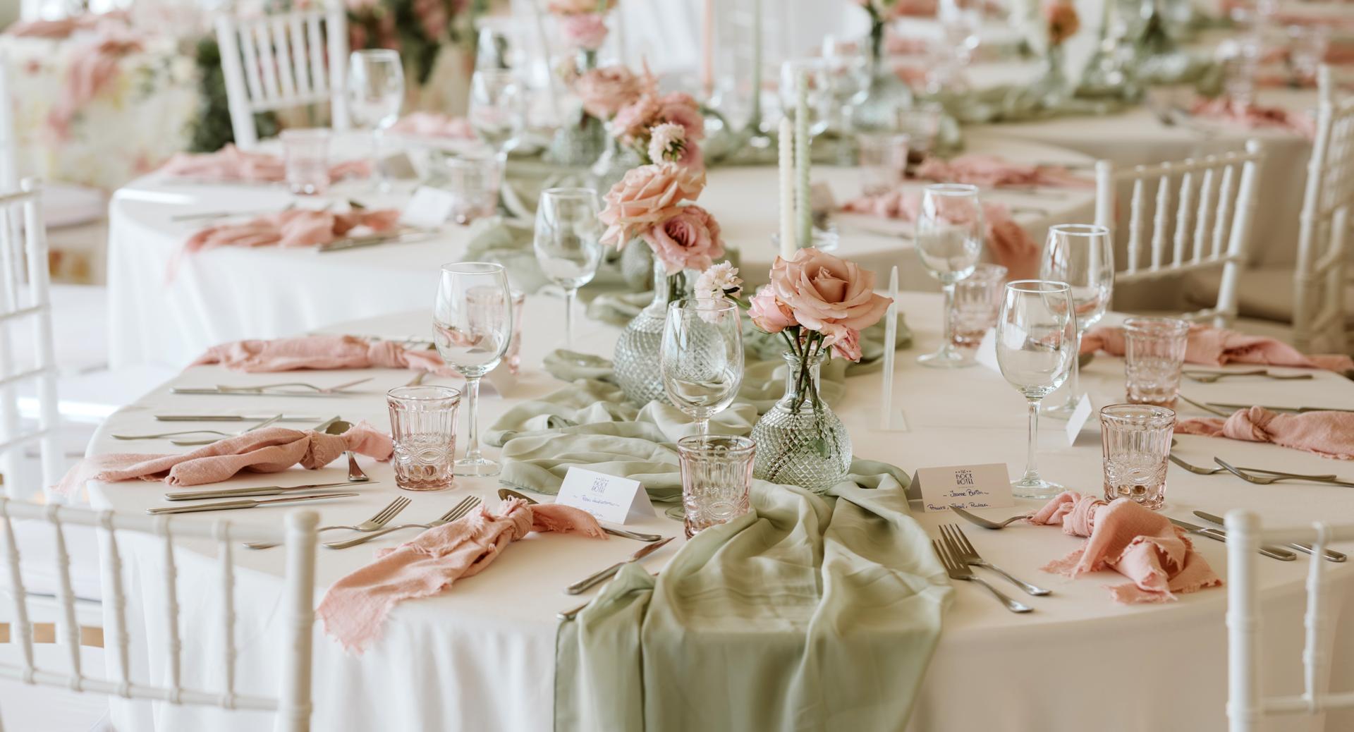 Round tables set for an event with glassware, cutlery, sage green table runners, pink napkins, and floral centerpieces with roses. White chairs surround the tables.