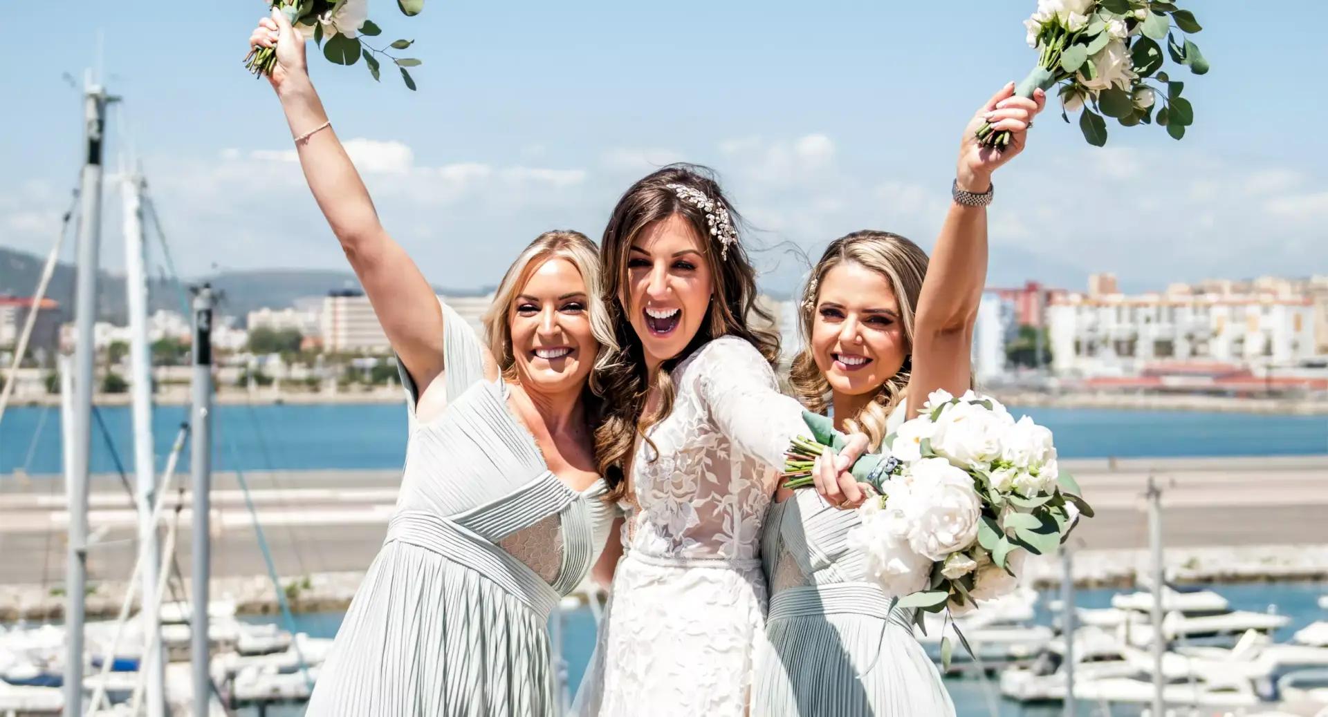 Three women in formal dresses pose joyfully outdoors with bouquets, with a marina, boats, and city buildings in the background.