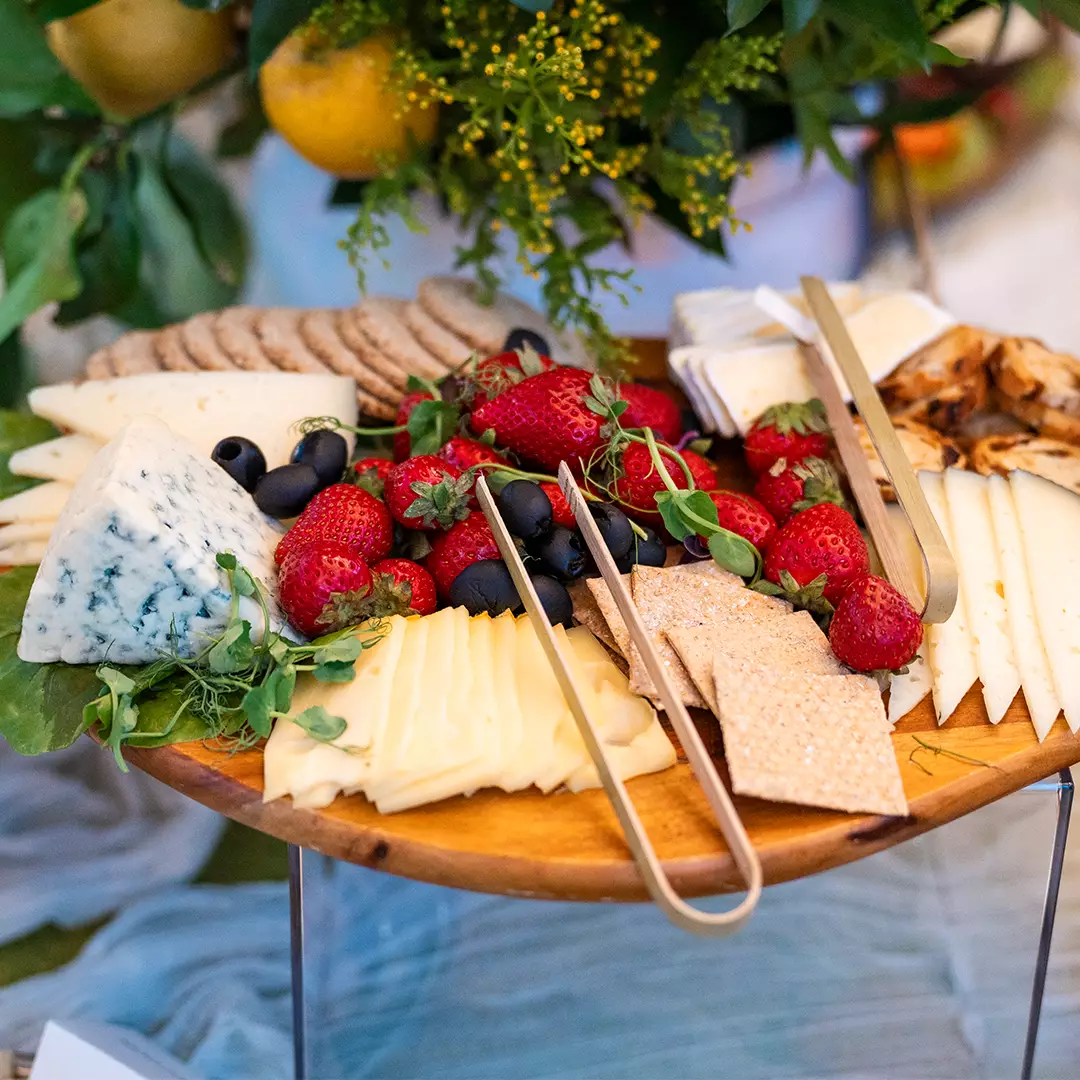 A wooden platter with assorted cheeses, crackers, strawberries, blueberries, and serving tongs, set on a table with greenery in the background.