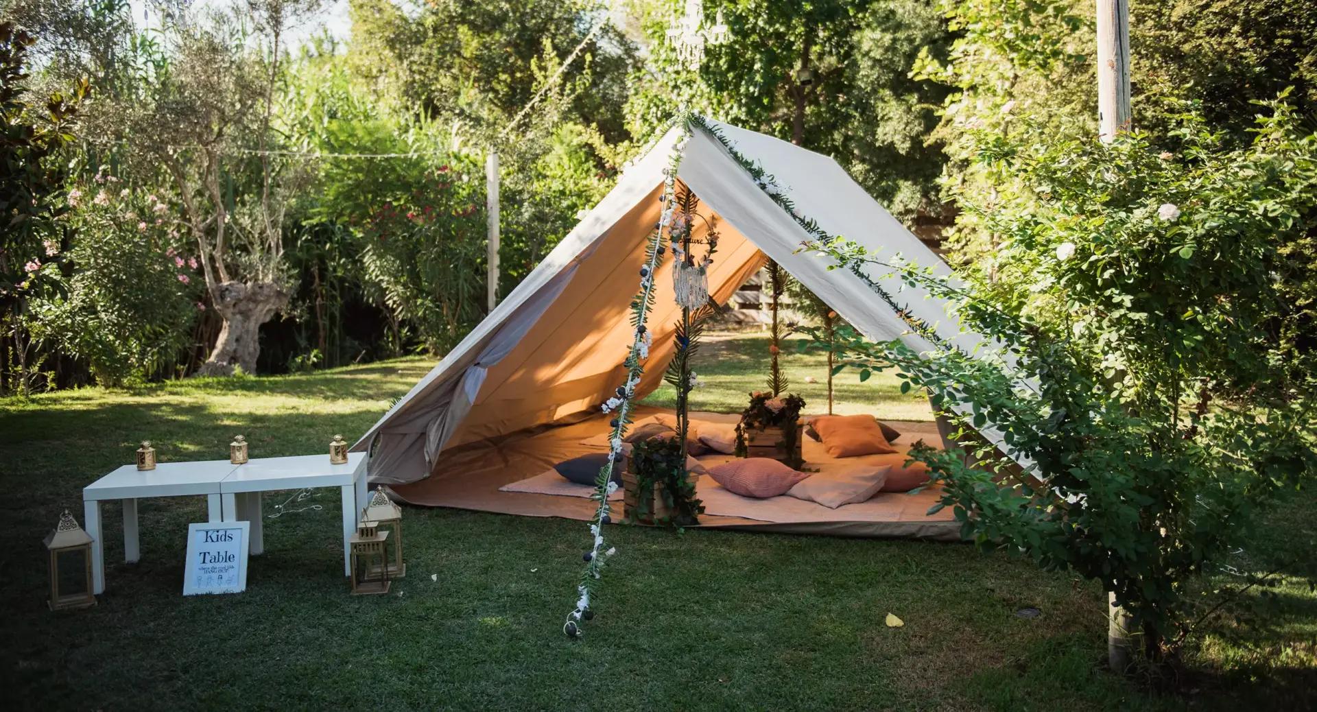 A beige tent with pillows inside is set up on a grassy lawn, surrounded by trees and greenery. A white table and lanterns are placed nearby.