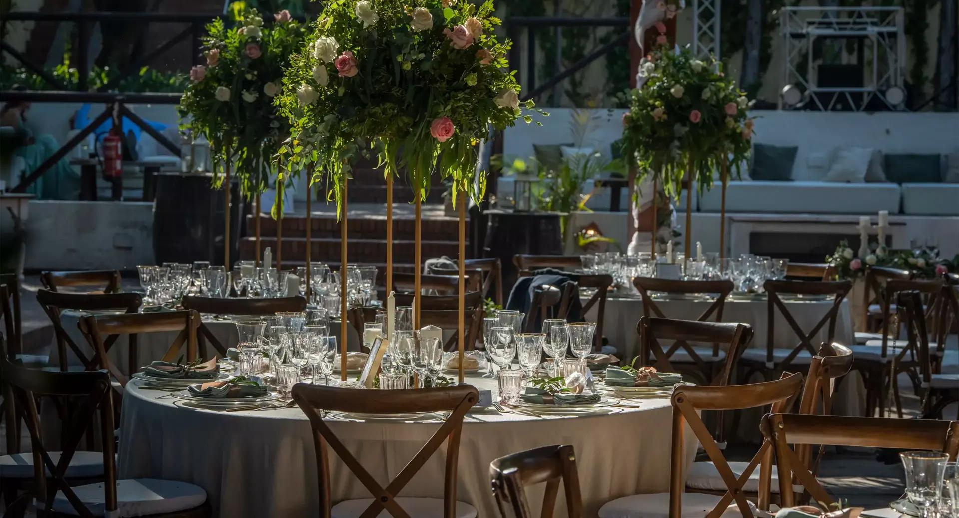Round table set for an event with glassware, plates, and floral centerpieces; wooden chairs surround the table in an outdoor or semi-outdoor venue.
