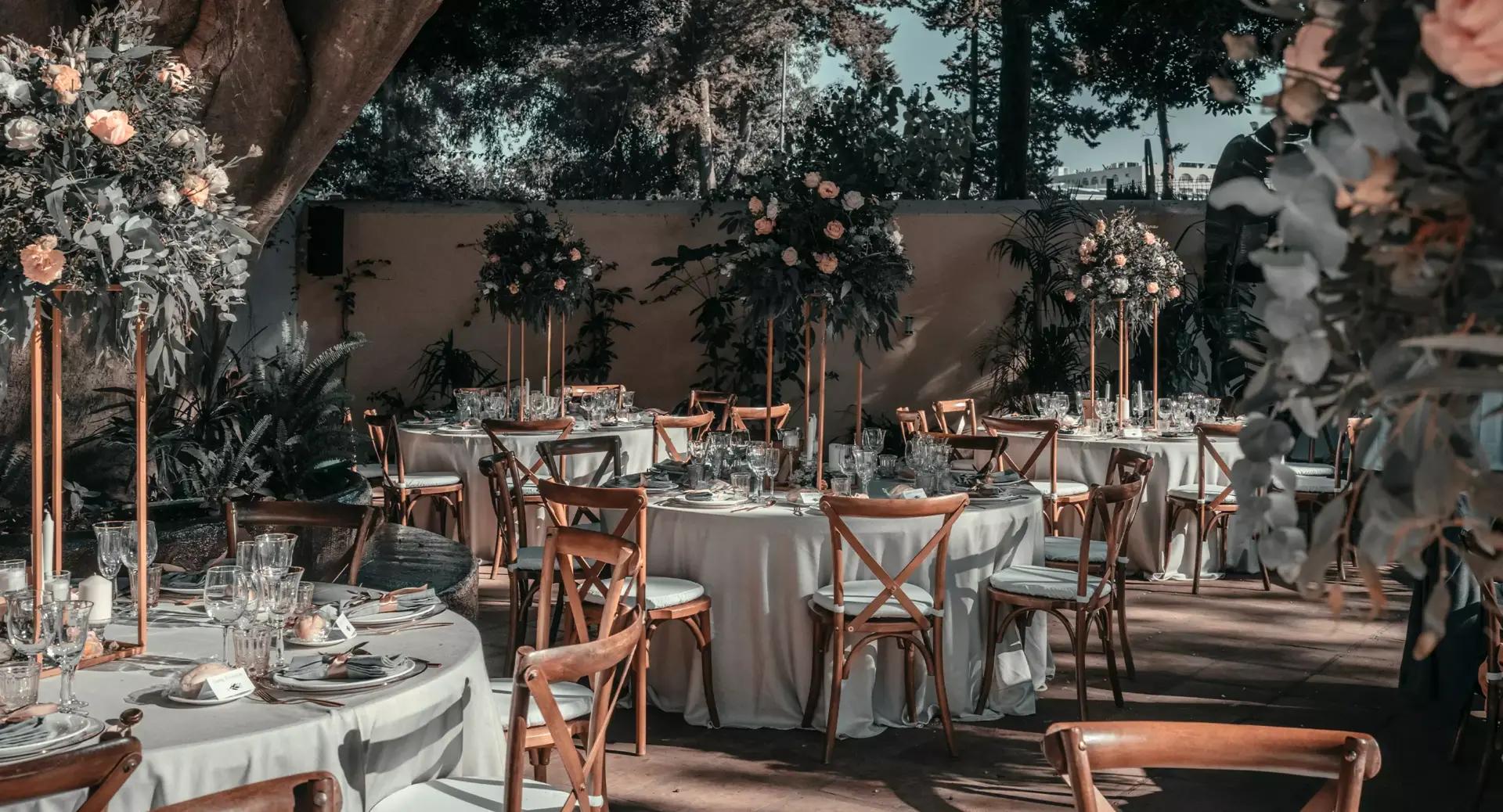 Outdoor event setup with round tables covered in white tablecloths, wooden chairs, floral centerpieces, glassware, and place settings under trees and natural light.