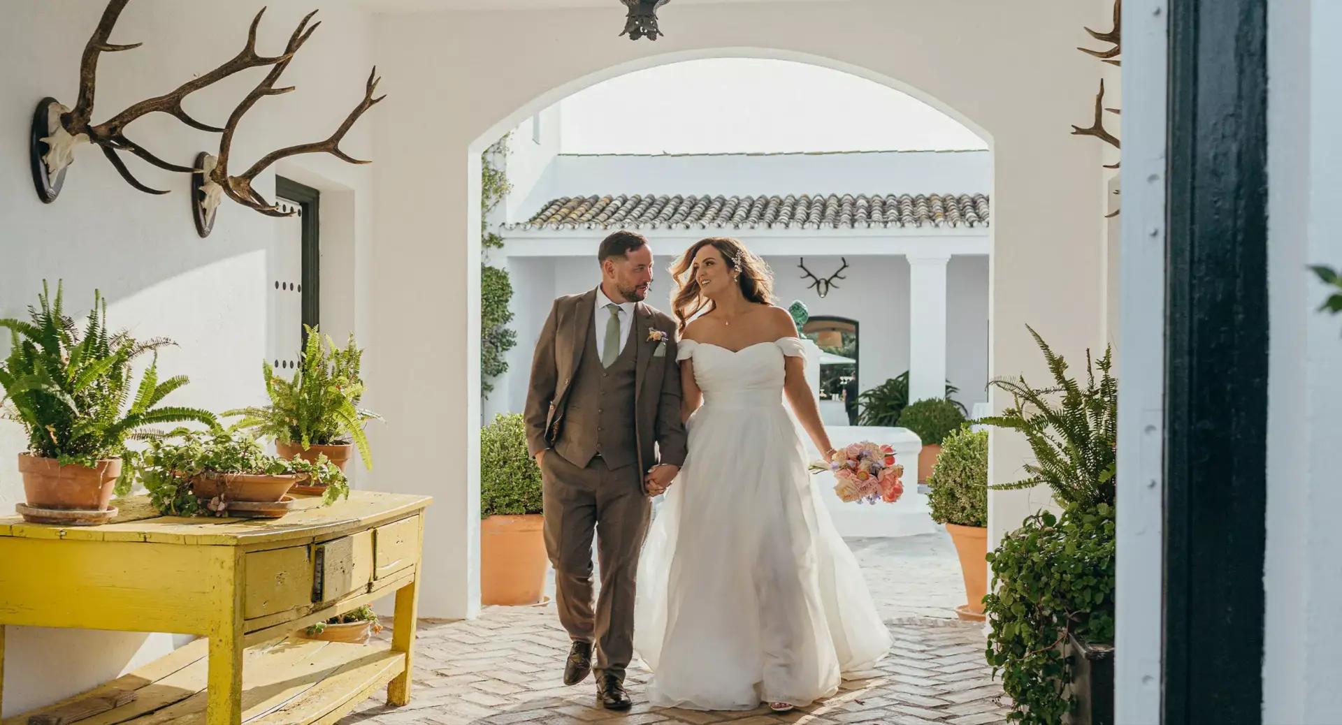 A bride and groom walk hand in hand through an arched outdoor corridor, surrounded by potted plants and antler decorations, with sunlight illuminating the scene.