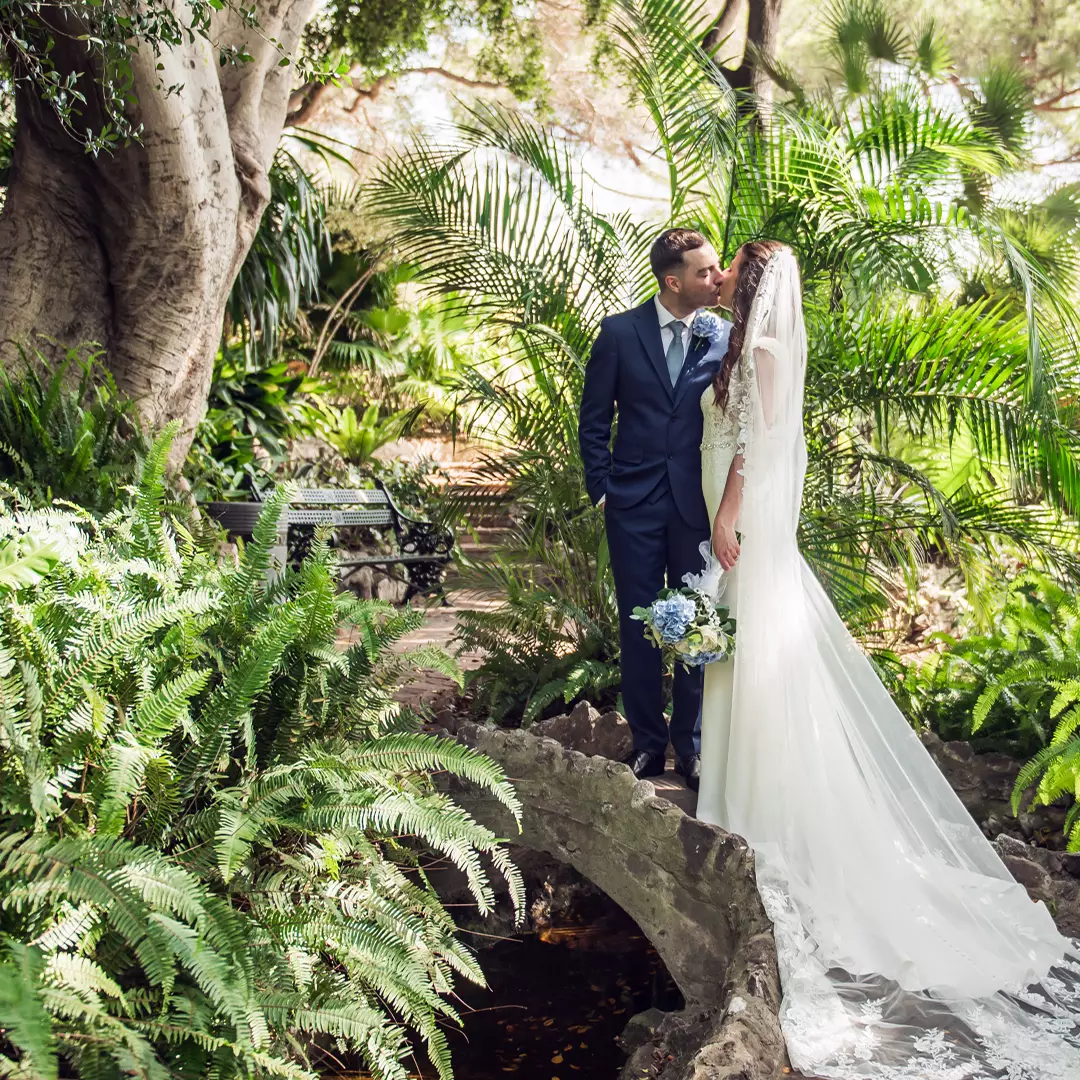 A bride and groom stand on a stone bridge in a lush garden, facing each other and kissing. The bride holds a bouquet and wears a long white dress with a veil.