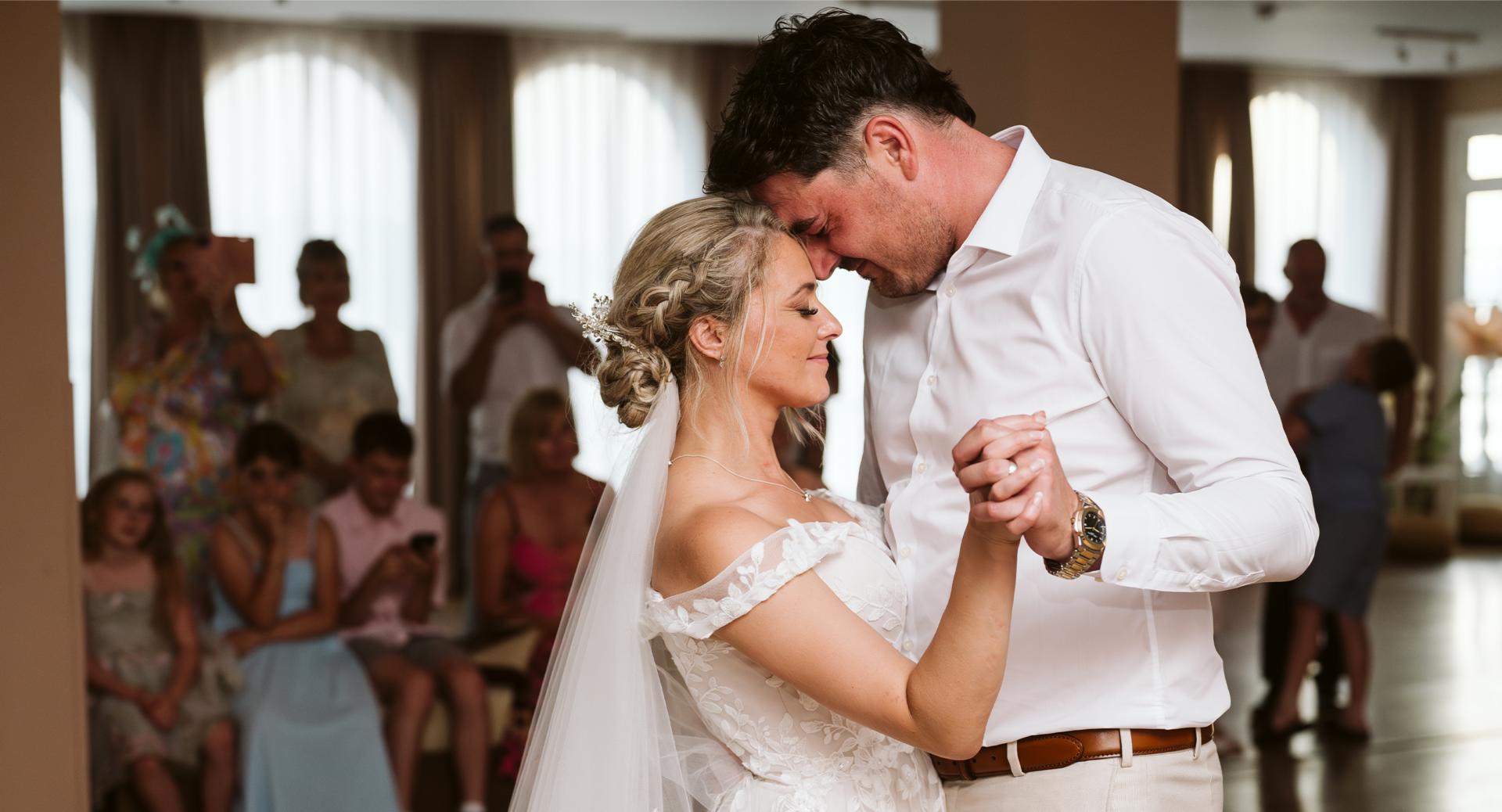 A bride and groom share a dance at their wedding reception, holding hands and touching foreheads, with guests seated and watching in the background.