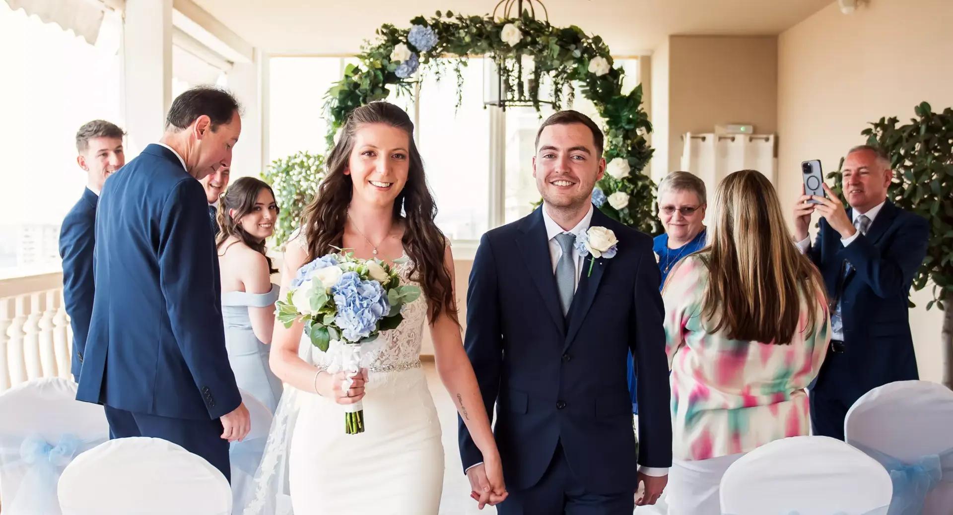 A bride and groom walk down the aisle holding hands, smiling, with family and friends standing and taking photos in a decorated indoor venue.