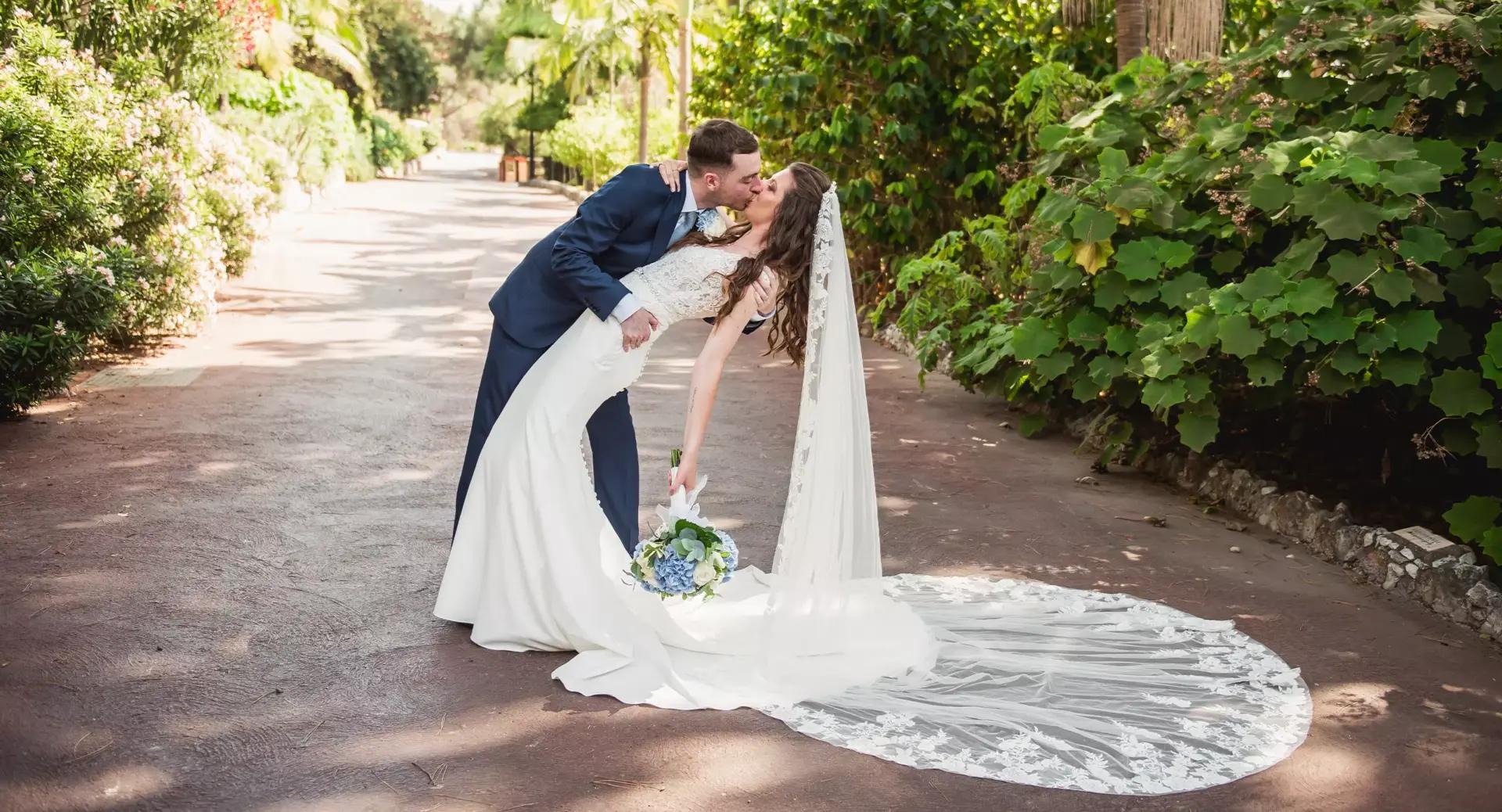 A bride in a white gown and veil holding a bouquet is dipped and kissed by a groom in a suit on an outdoor path surrounded by greenery.