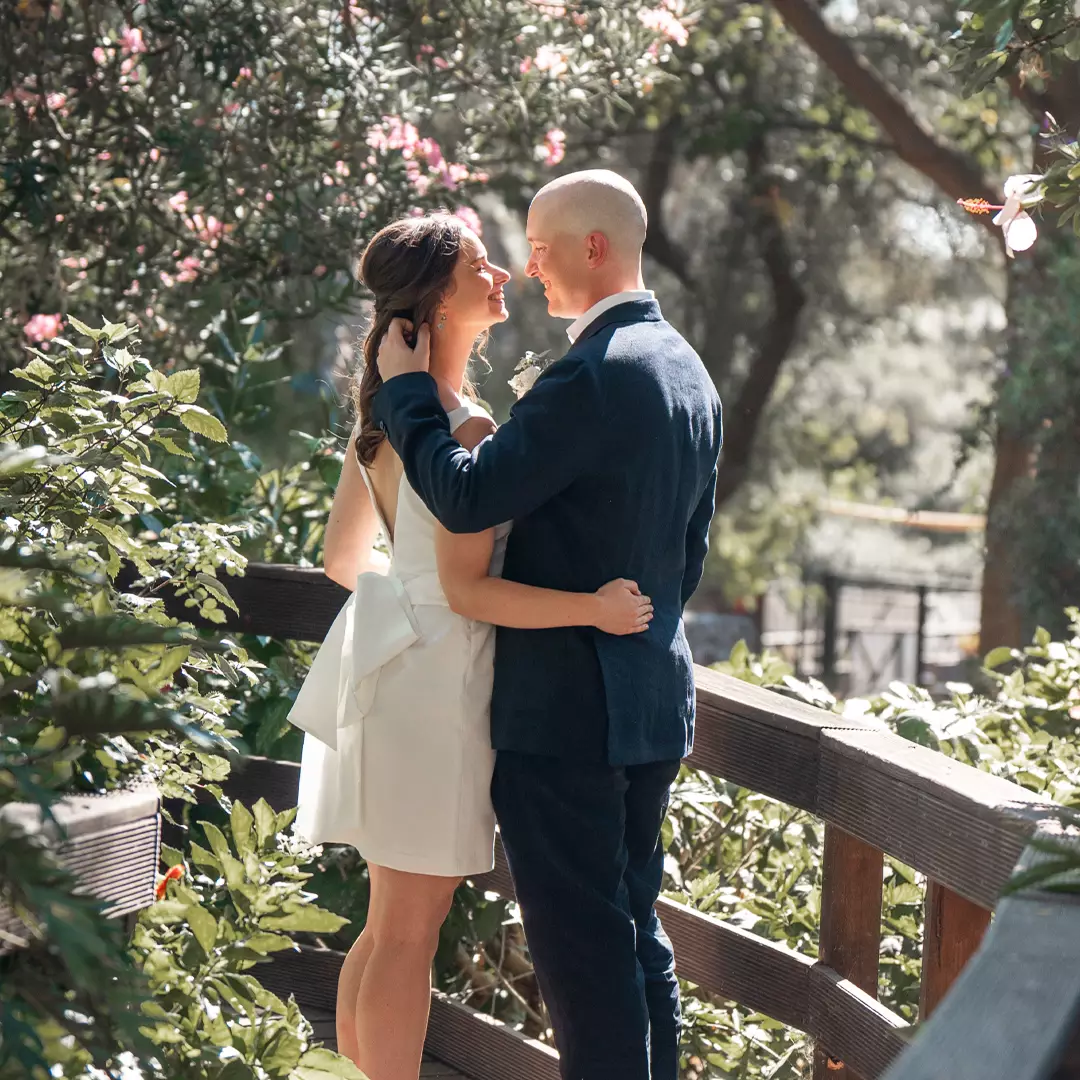 A couple stands on a wooden bridge surrounded by greenery, facing each other and embracing in sunlight.