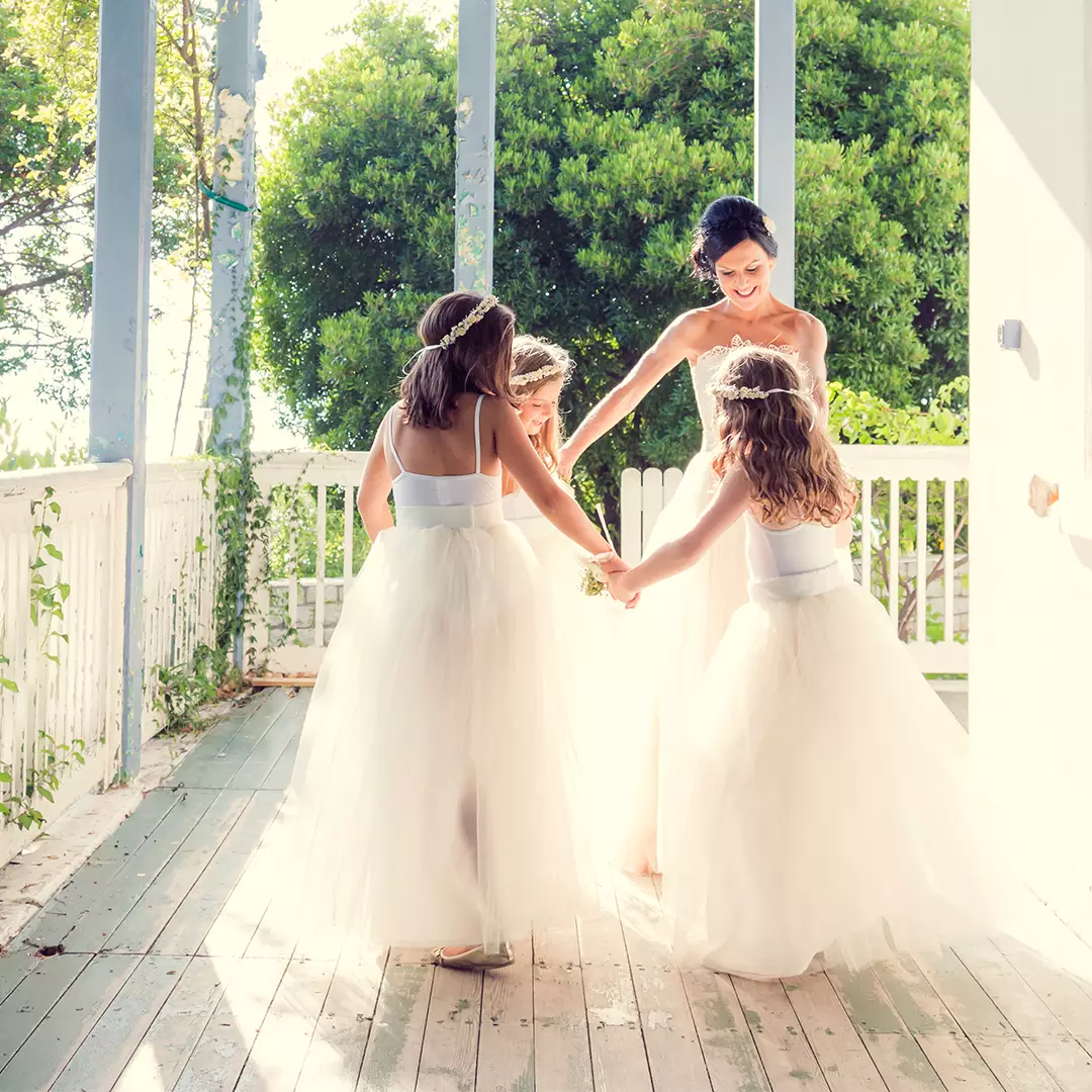 A bride in a white gown holds hands in a circle with three young girls in matching white dresses on a sunlit porch.