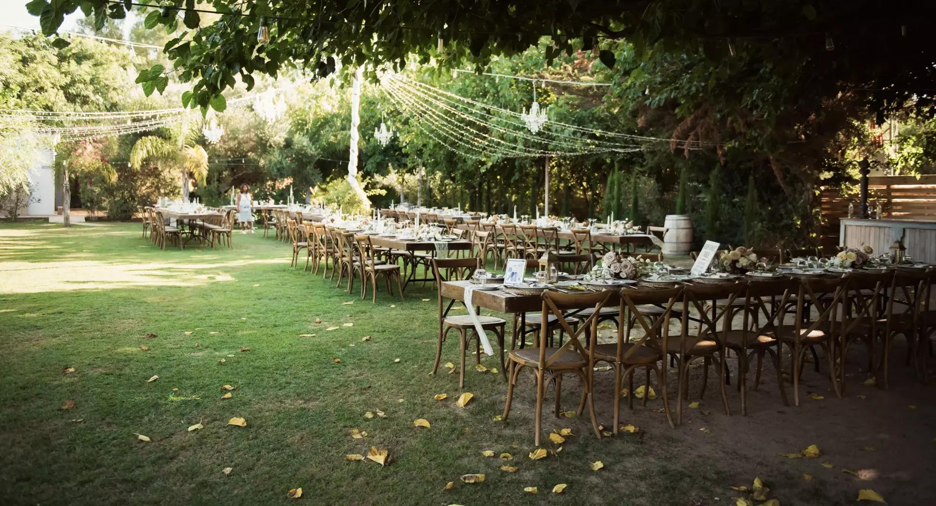 Outdoor event setup with long tables, wooden chairs, floral centerpieces, and string lights under trees on a grassy lawn.