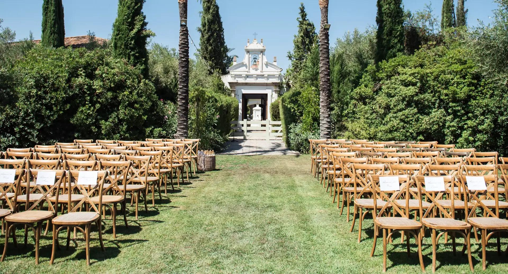 Rows of wooden chairs are arranged on a grassy lawn between tall trees, facing a small white building, set up for an outdoor ceremony or event.