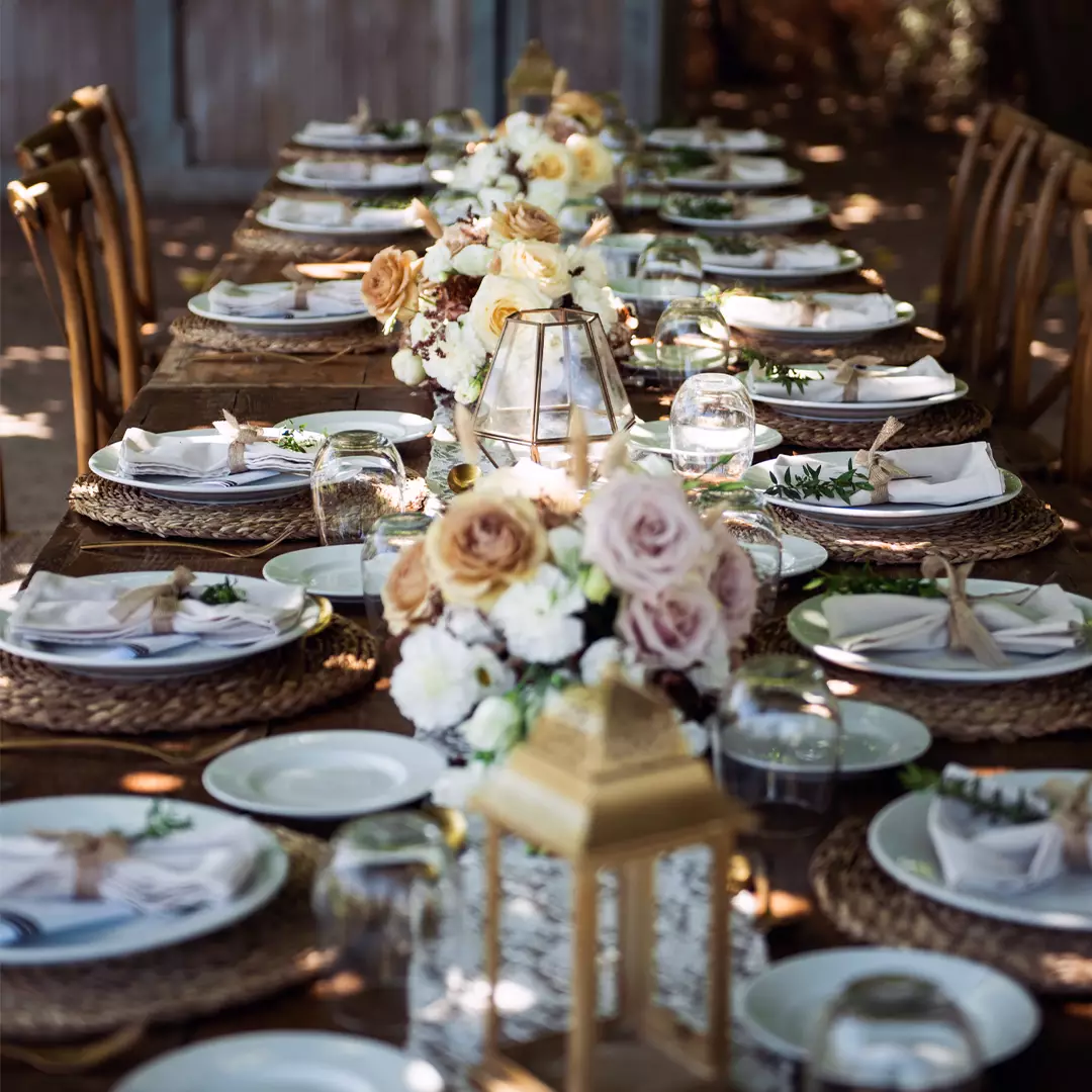 A long wooden table set for a formal meal with plates, napkins, glasses, flowers, and lantern centerpieces arranged neatly.