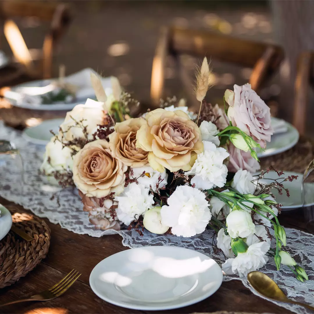 A floral centerpiece with beige and white roses, greenery, and lace decorates a rustic wooden table set with white plates and woven placemats.