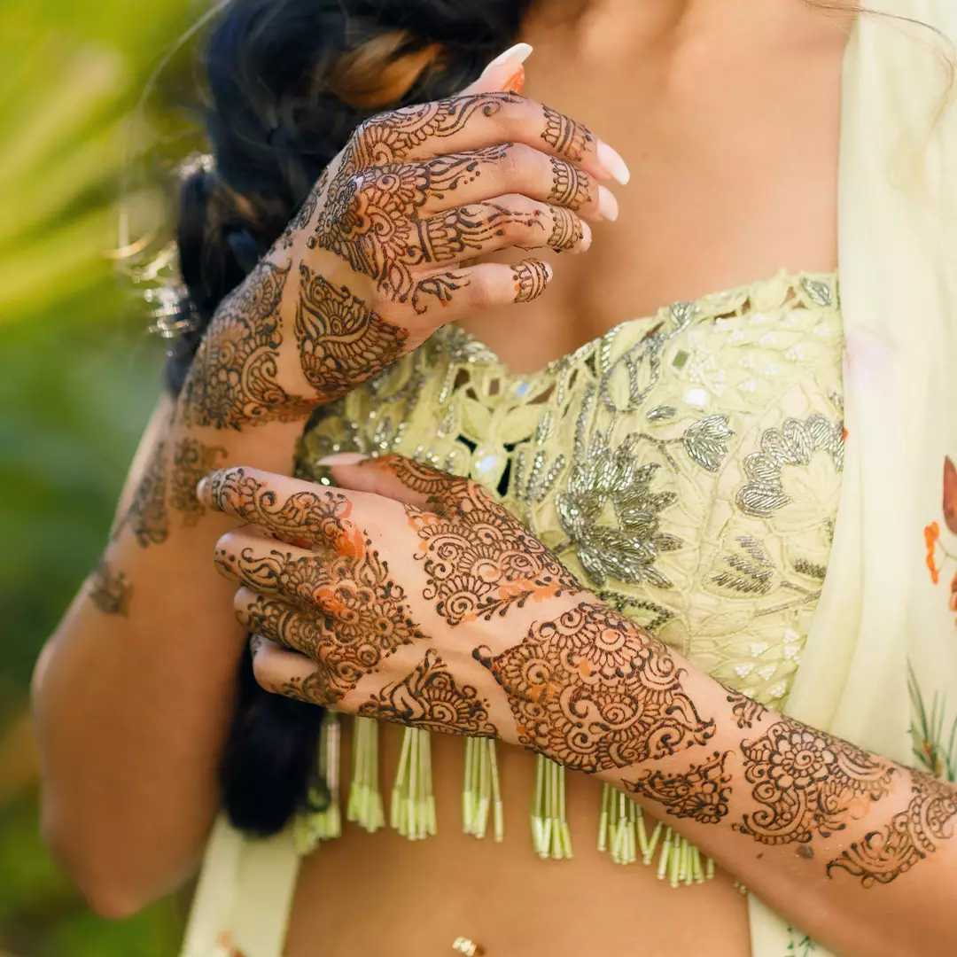 A woman wearing a light-colored, embellished outfit displays intricate henna designs on both hands and forearms.