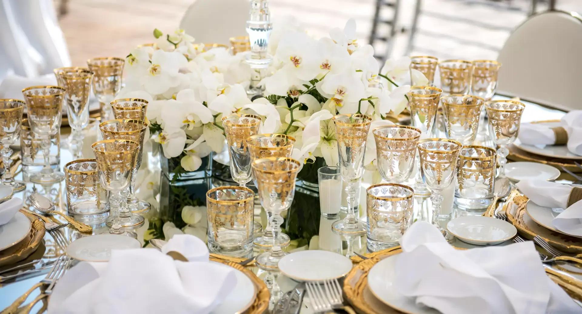 Formal table setting with gold-trimmed glassware, white napkins, and a centerpiece of white orchids arranged on a mirrored surface.