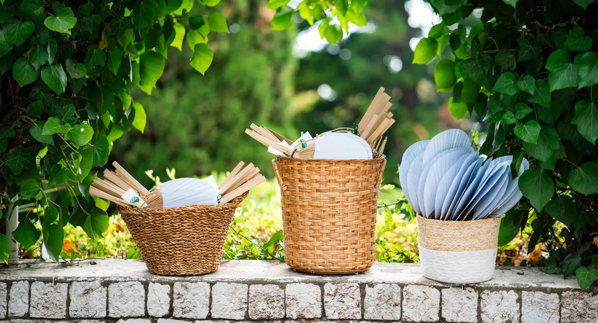 Three woven baskets sit on a stone ledge outdoors, each filled with handheld paper fans and folding fans, surrounded by green foliage.