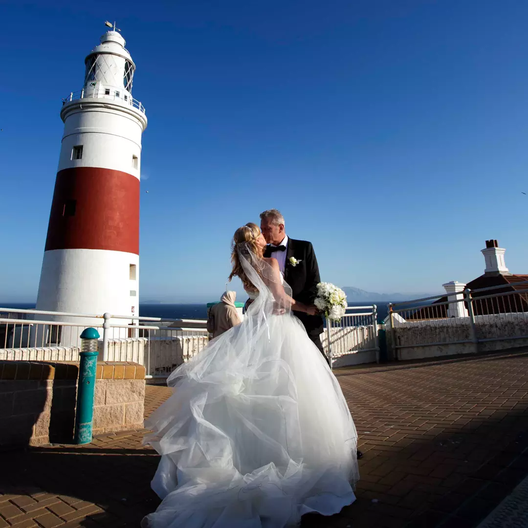 A bride and groom kiss in front of a white and red lighthouse under a clear blue sky.