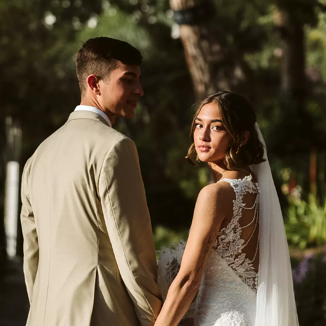 A bride in a white lace dress with a veil and a groom in a beige suit stand outdoors, holding hands and looking back at the camera.