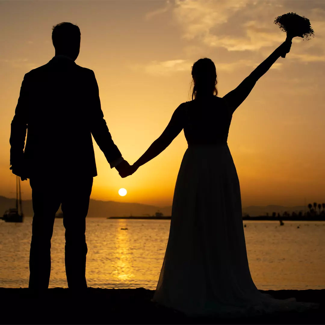 Silhouetted couple holding hands by the water at sunset, with one person raising a bouquet in the air.
