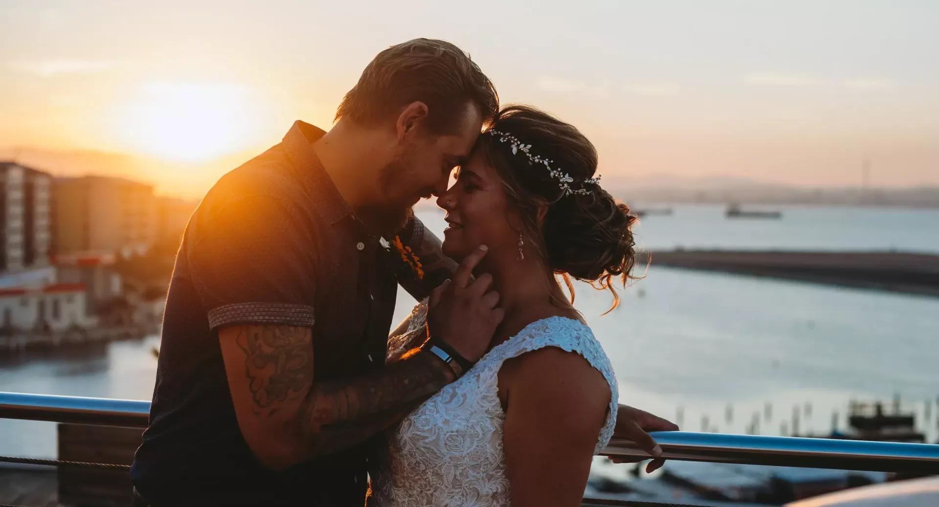 A couple embraces and smiles at each other on a balcony at sunset, with water and buildings visible in the background.