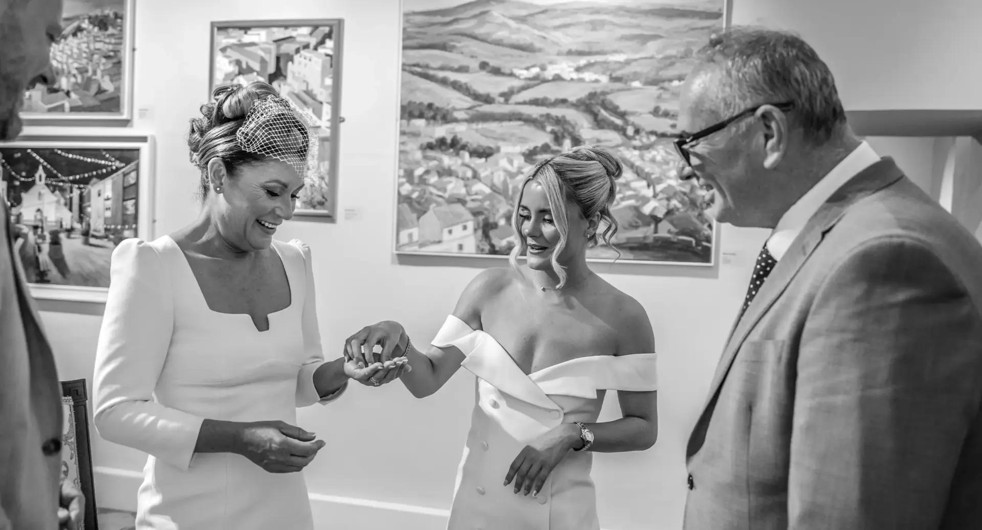 Two women in formal dresses exchange rings and smile, while two men in suits stand nearby, inside an art gallery with paintings on the wall.