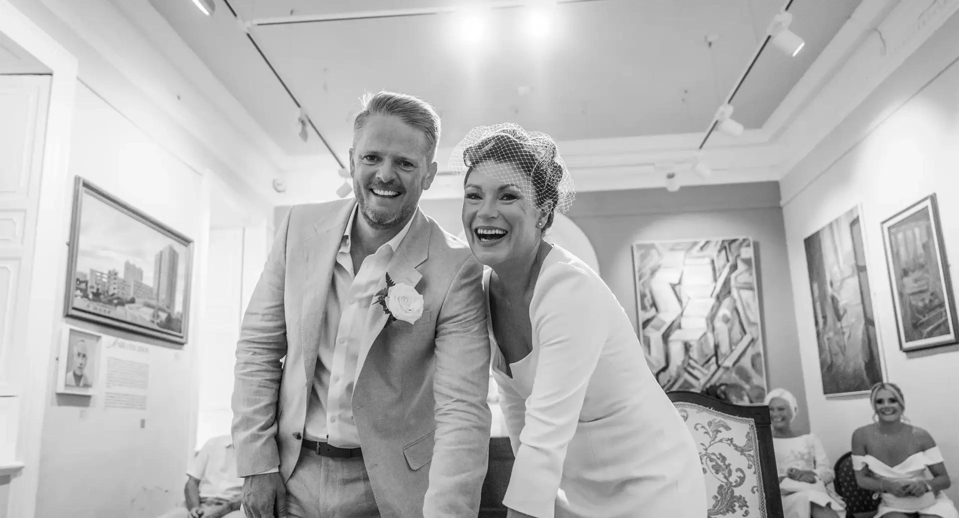 A smiling couple in wedding attire leans forward together in an art gallery, surrounded by paintings and seated guests.