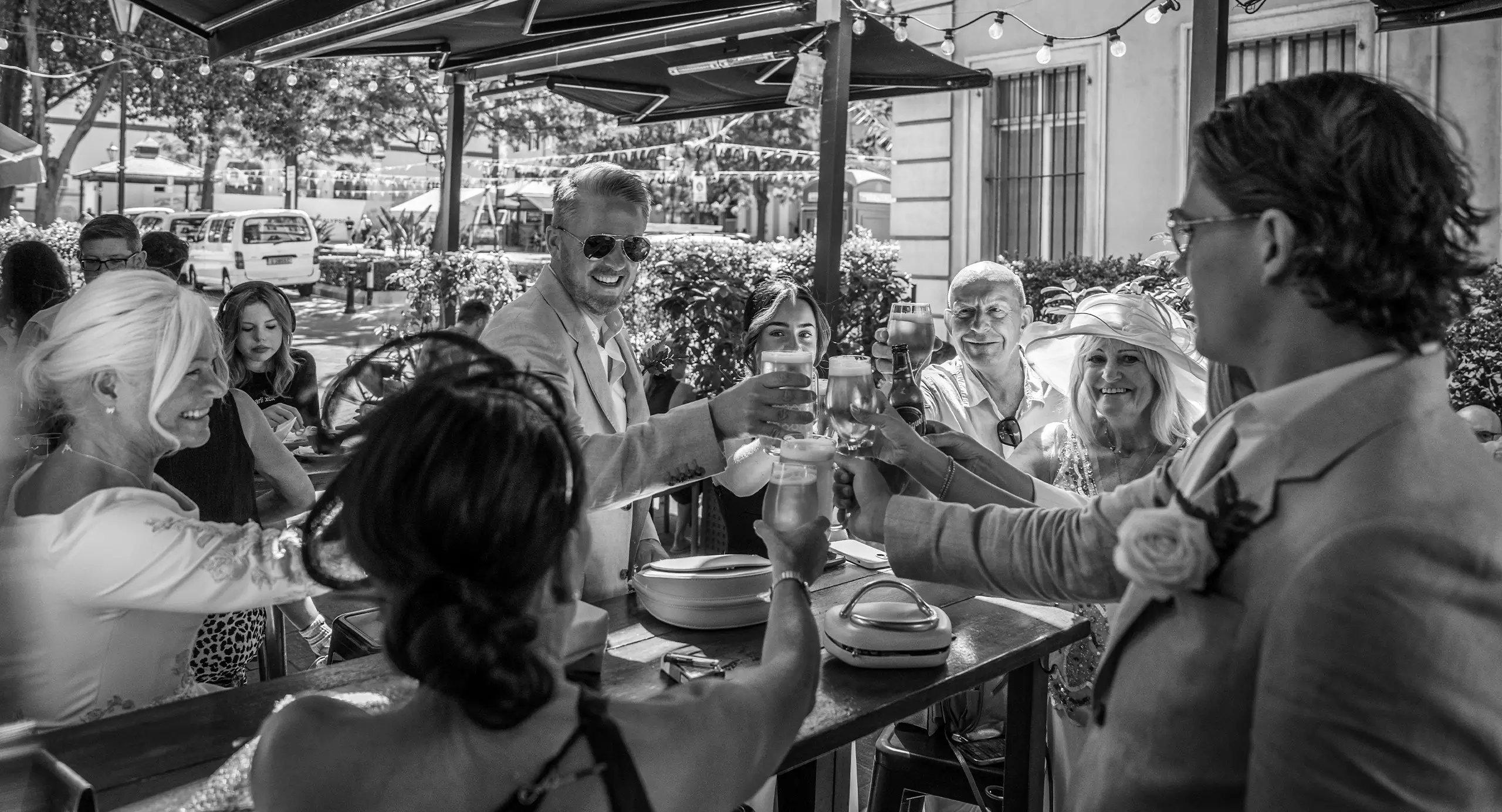 A group of people dressed in formal attire are gathered outdoors, raising glasses in a toast around a table under umbrellas.