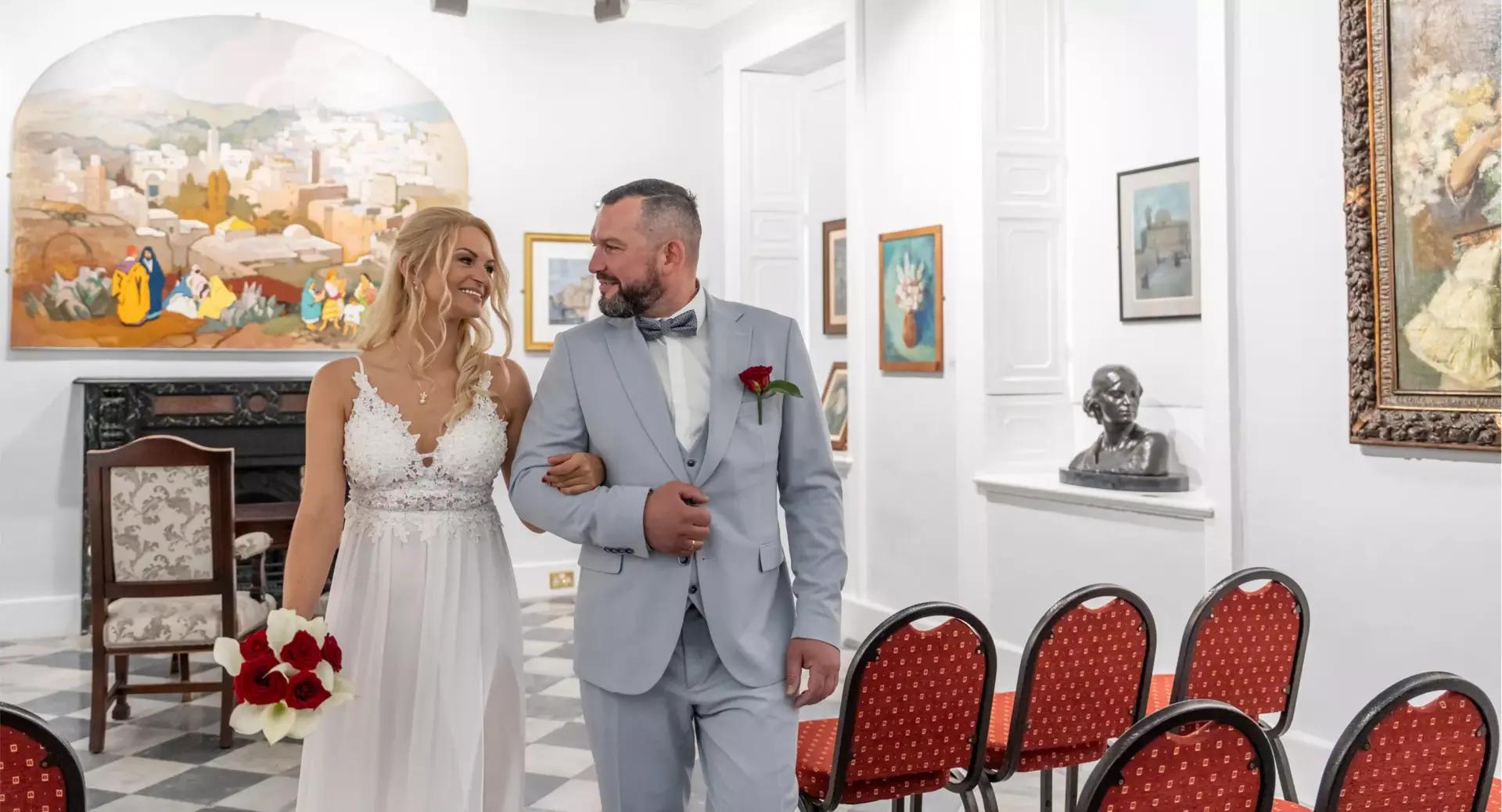 A bride and groom walk arm in arm down an aisle in a small, art-filled room with red chairs and framed paintings on the walls.