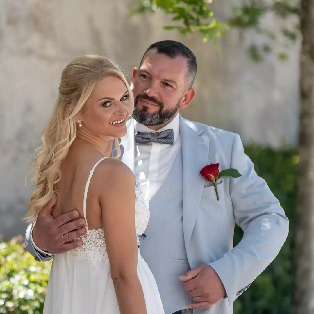 A bride in a white dress and a groom in a light gray suit with a red rose stand outdoors, looking at the camera and smiling.