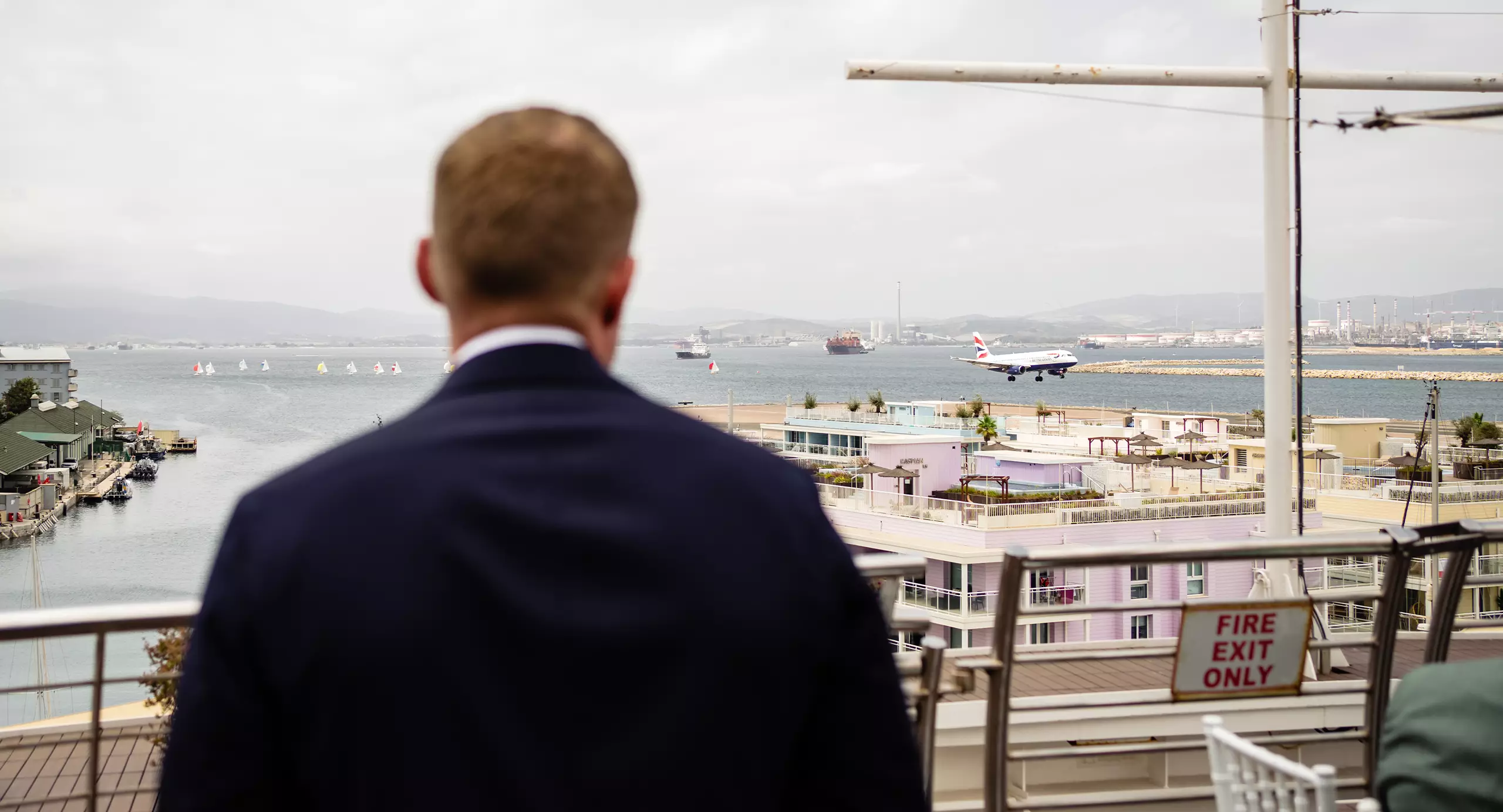 A man in a suit stands on a balcony overlooking a harbor with boats and a plane landing on a runway in the distance.