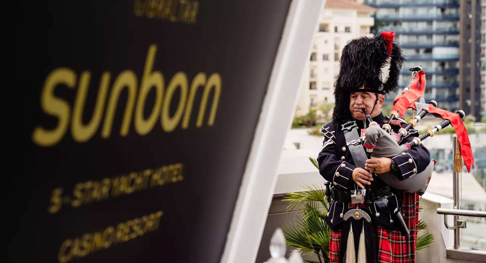 A bagpiper in traditional attire plays near a sign for the Sunborn hotel, with buildings visible in the background.