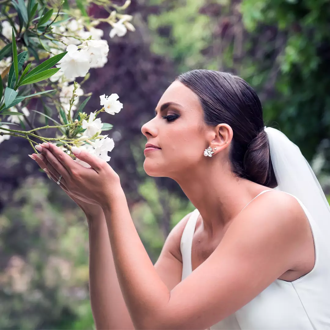 A bride in a white dress with a veil smells white flowers outdoors, surrounded by greenery.