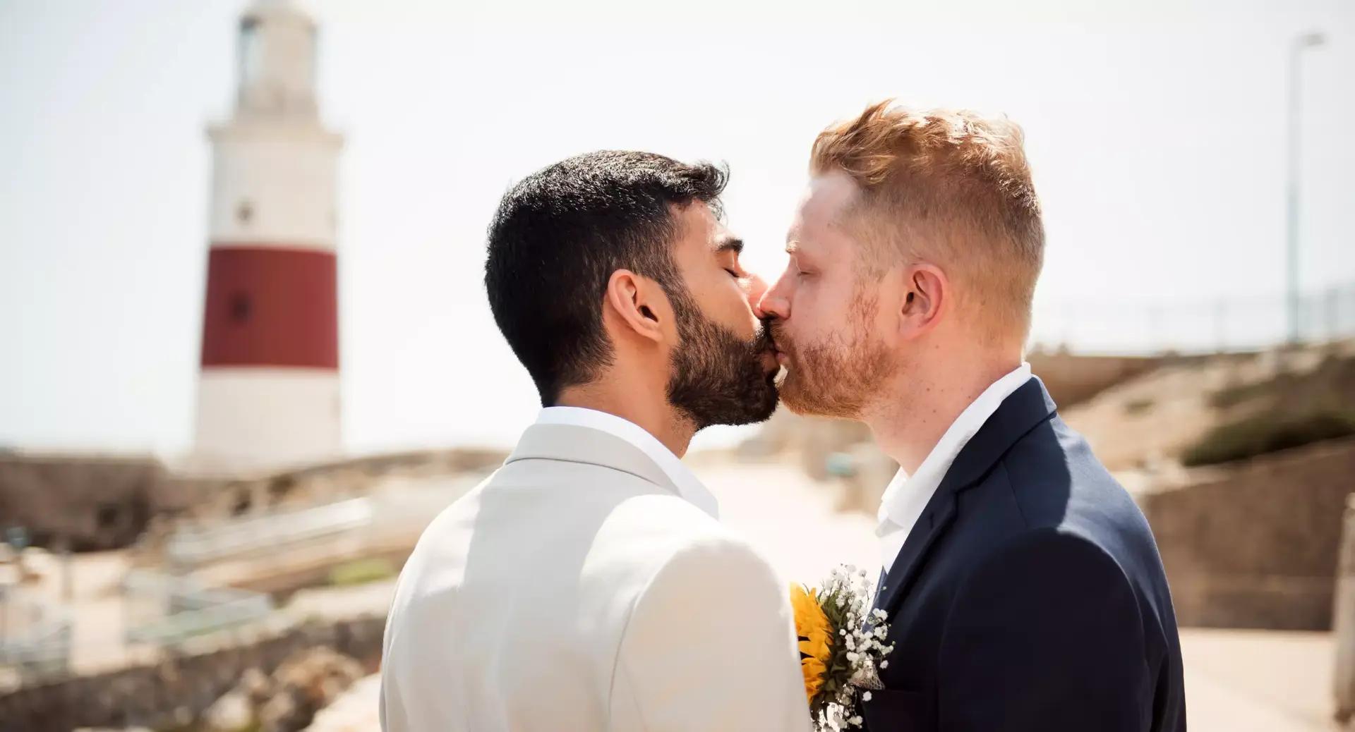 Two men in suits kiss outdoors near a lighthouse, one holding a sunflower bouquet.