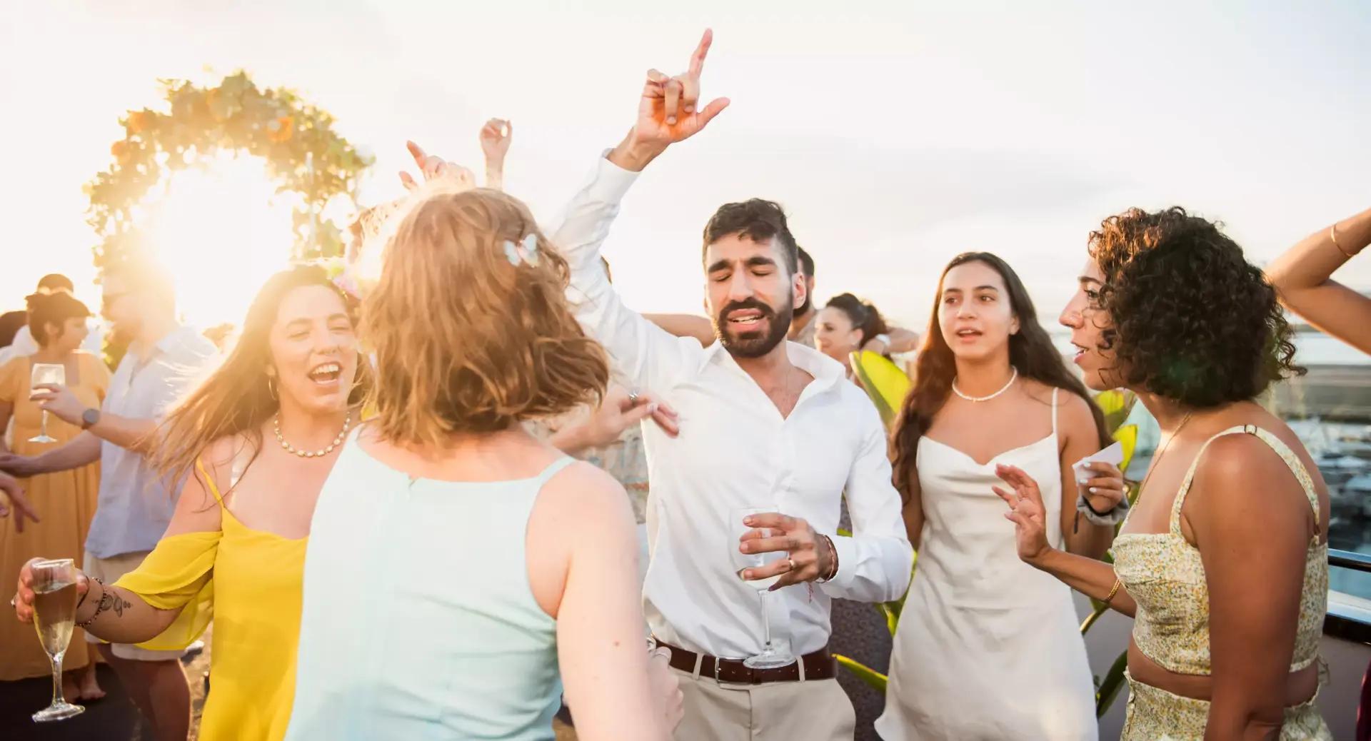 A group of people, dressed in semi-formal attire, dance and celebrate outdoors in daylight, with drinks in hand and the sun shining in the background.