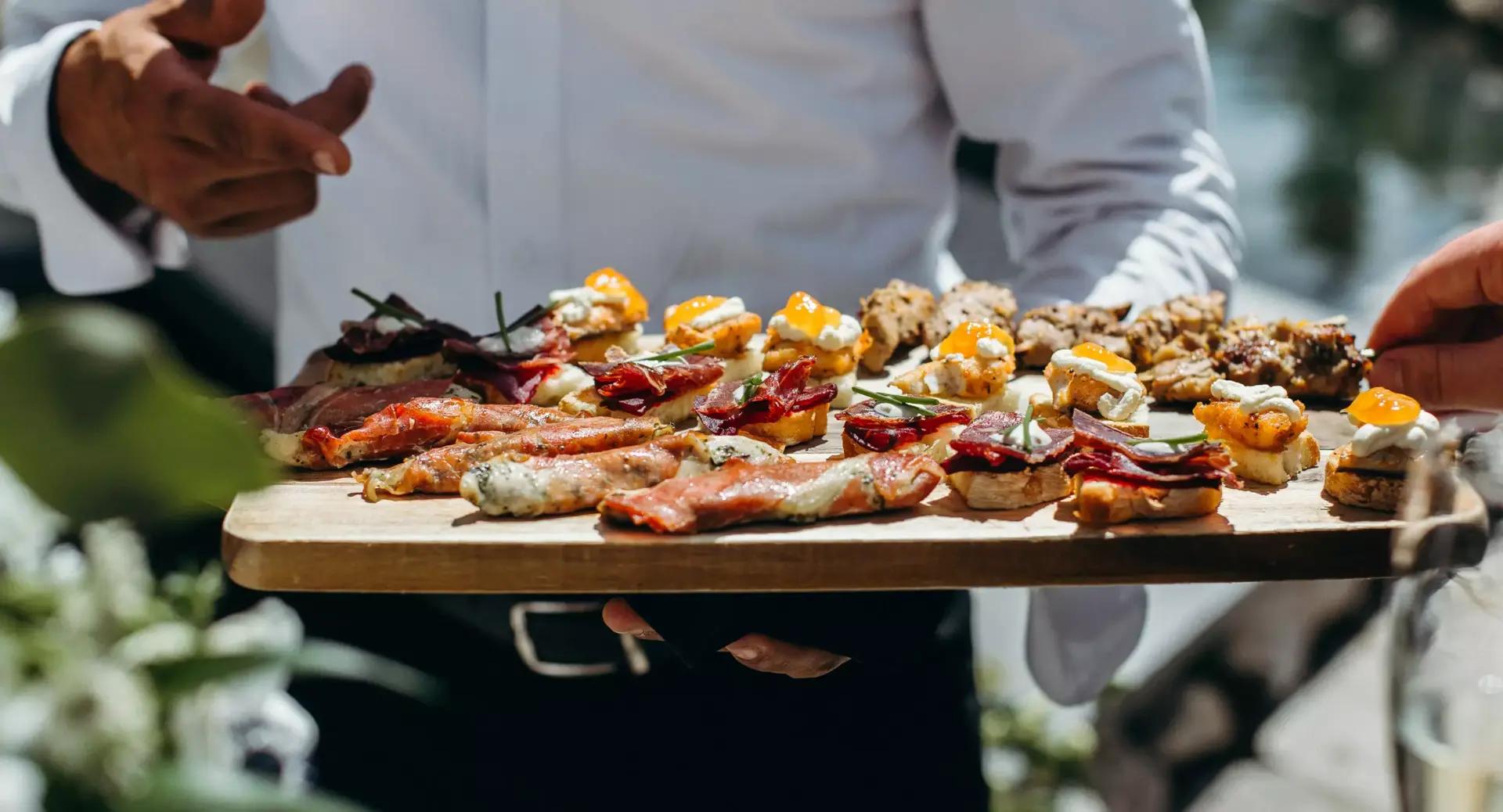 A person in a white shirt holds a wooden platter with assorted appetizers, including meats, cheeses, and garnished bites, while another hand reaches for food.