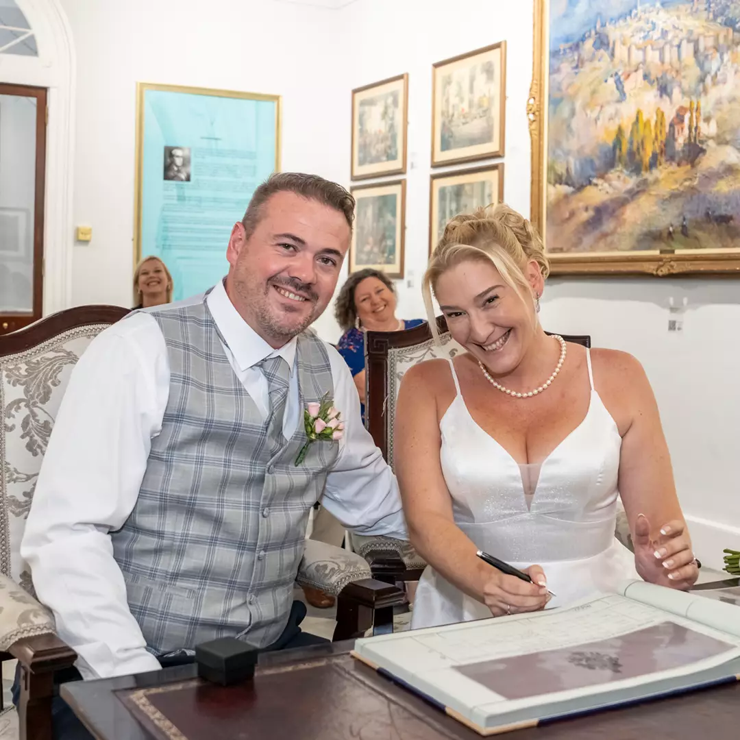 A bride and groom sit together, smiling, as the bride signs a document at a table during a wedding ceremony. Paintings and framed items are visible on the walls behind them.