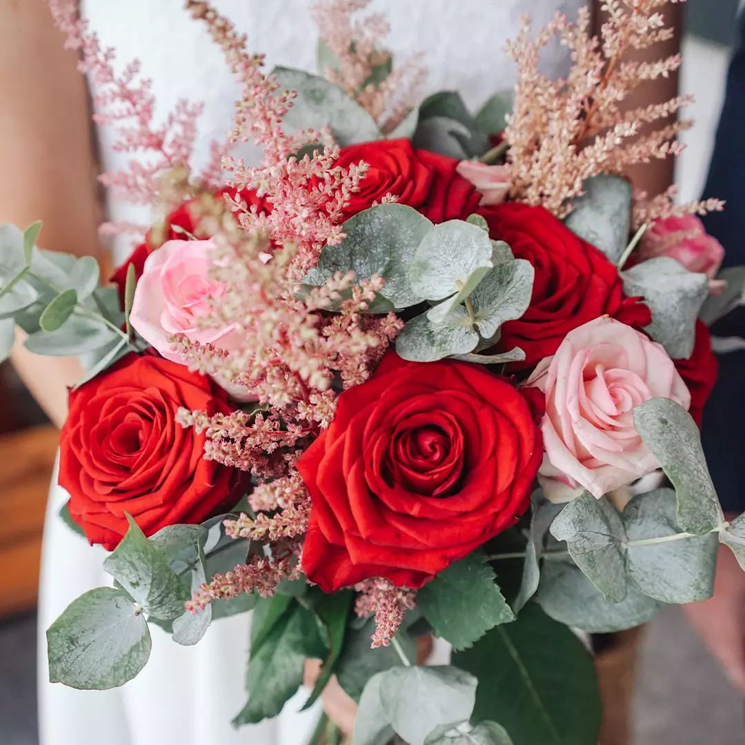 A close-up of a person holding a bouquet with red and pink roses, green leaves, and small pink filler flowers.