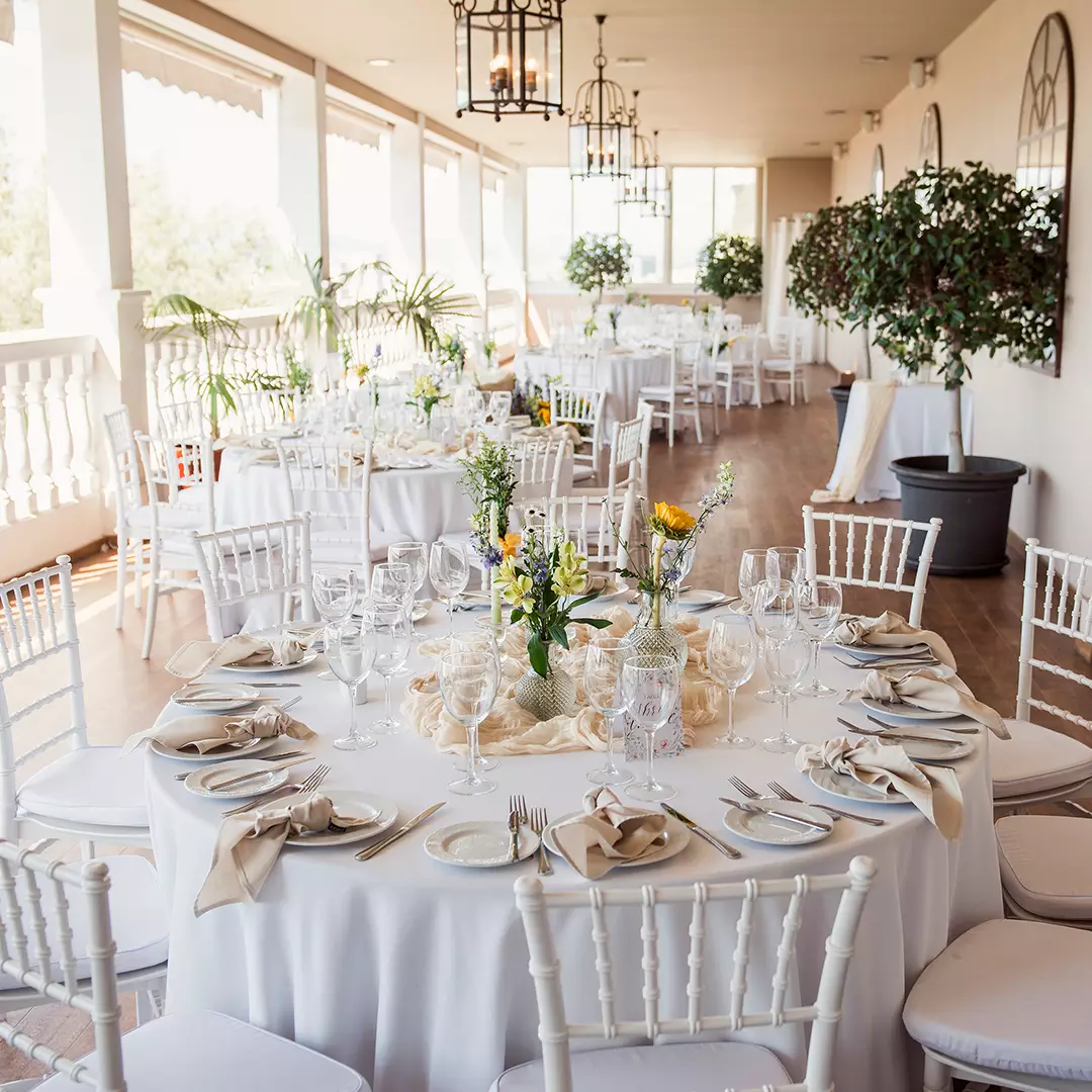 Elegant outdoor event space with round tables set with white linens, glassware, and floral centerpieces; white chairs and potted plants decorate the area.