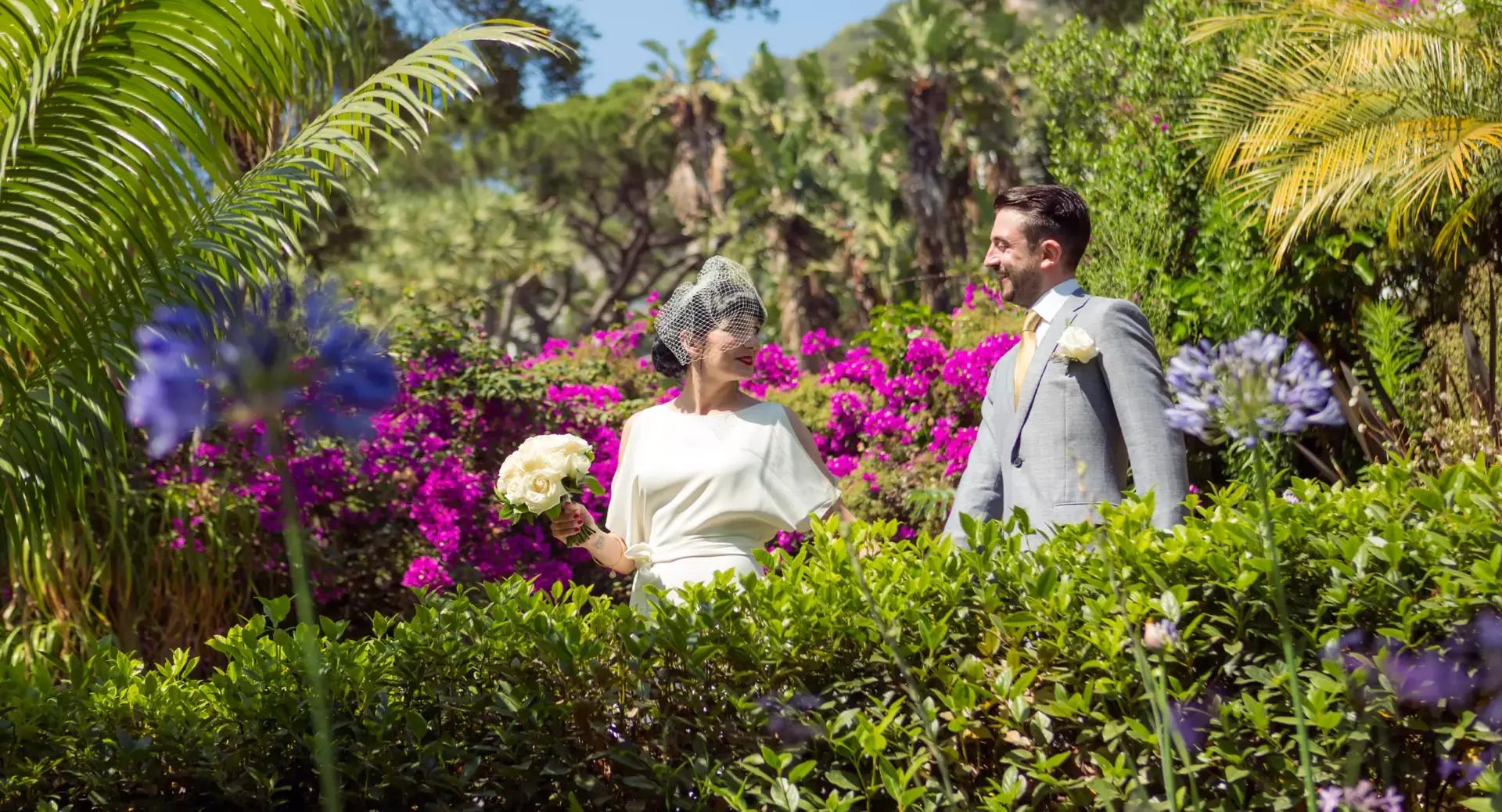 A bride and groom stand in a lush garden with vibrant pink flowers, greenery, and palm trees, smiling at each other on a sunny day.