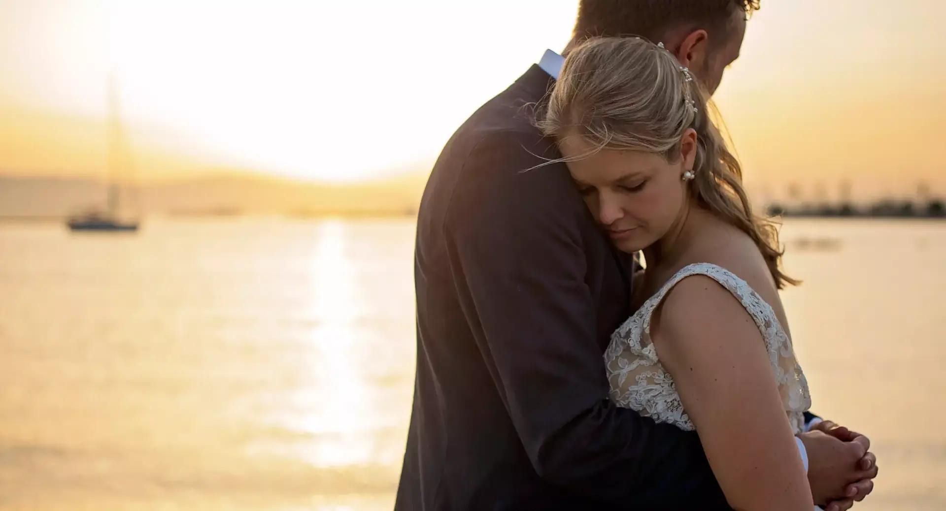 A couple in wedding attire embraces by the water at sunset, with a sailboat and soft sunlight in the background.