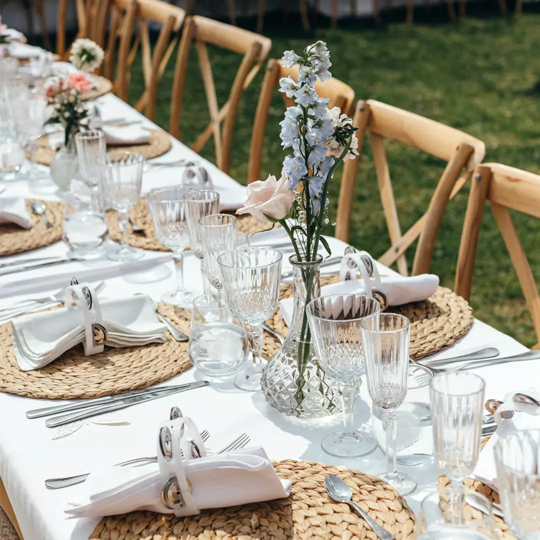 A long outdoor dining table set with woven placemats, glassware, white napkins in rings, and vases of pastel flowers on a white tablecloth.