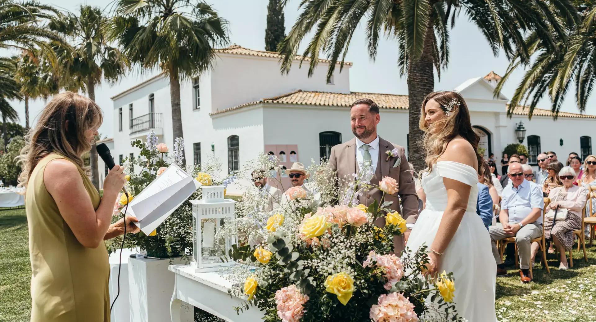 A couple stands at an outdoor wedding ceremony with a woman officiant speaking, surrounded by flowers, guests, and palm trees.