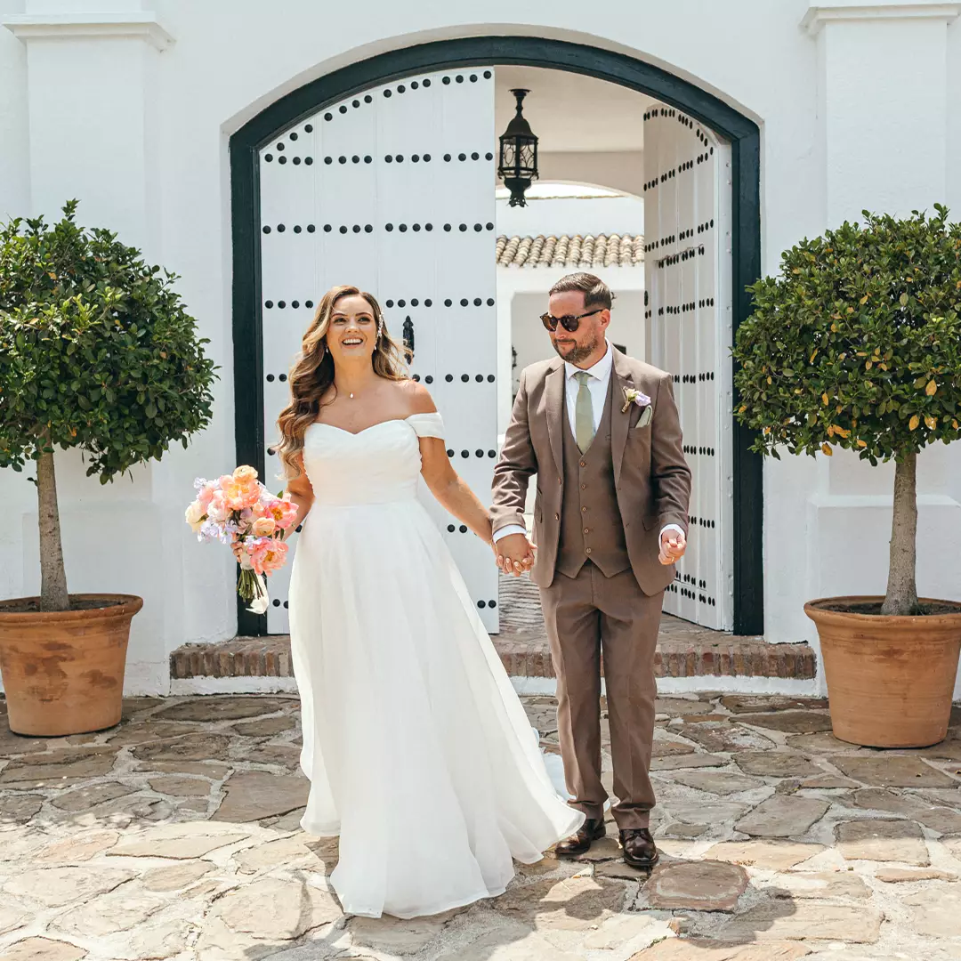 A bride in a white dress and a groom in a brown suit hold hands and smile while walking outside a white building with large potted trees.