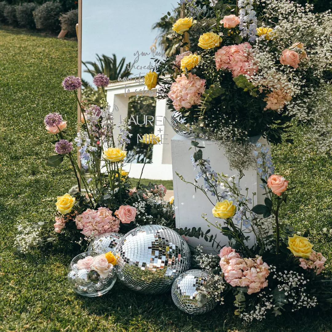 Floral arrangement with yellow and pink roses, baby's breath, and greenery next to a mirror and several disco balls on grass.