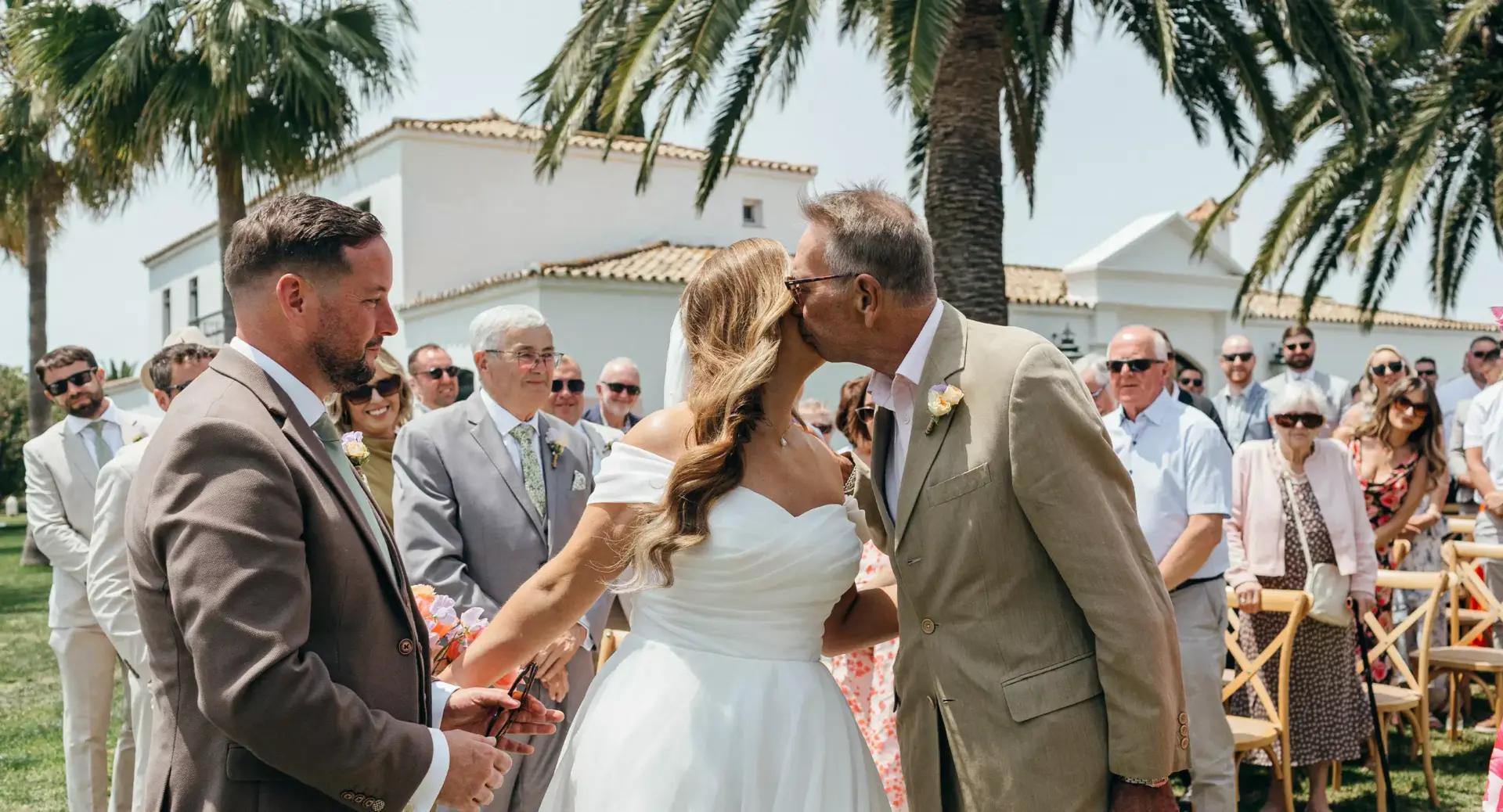 An older man kisses a bride on the cheek at an outdoor wedding ceremony, while a groom stands beside her and guests watch in the background.