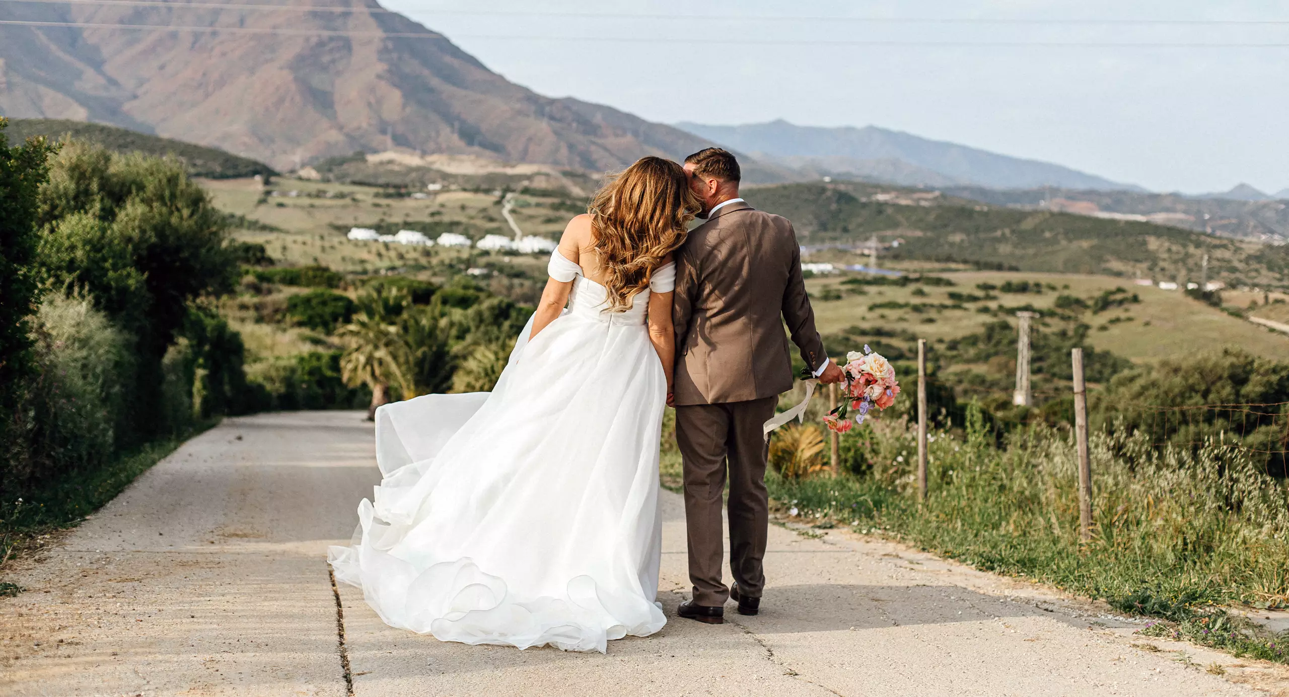 A bride in a white dress and a groom in a suit walk down a country road, with green hills and mountains in the background.