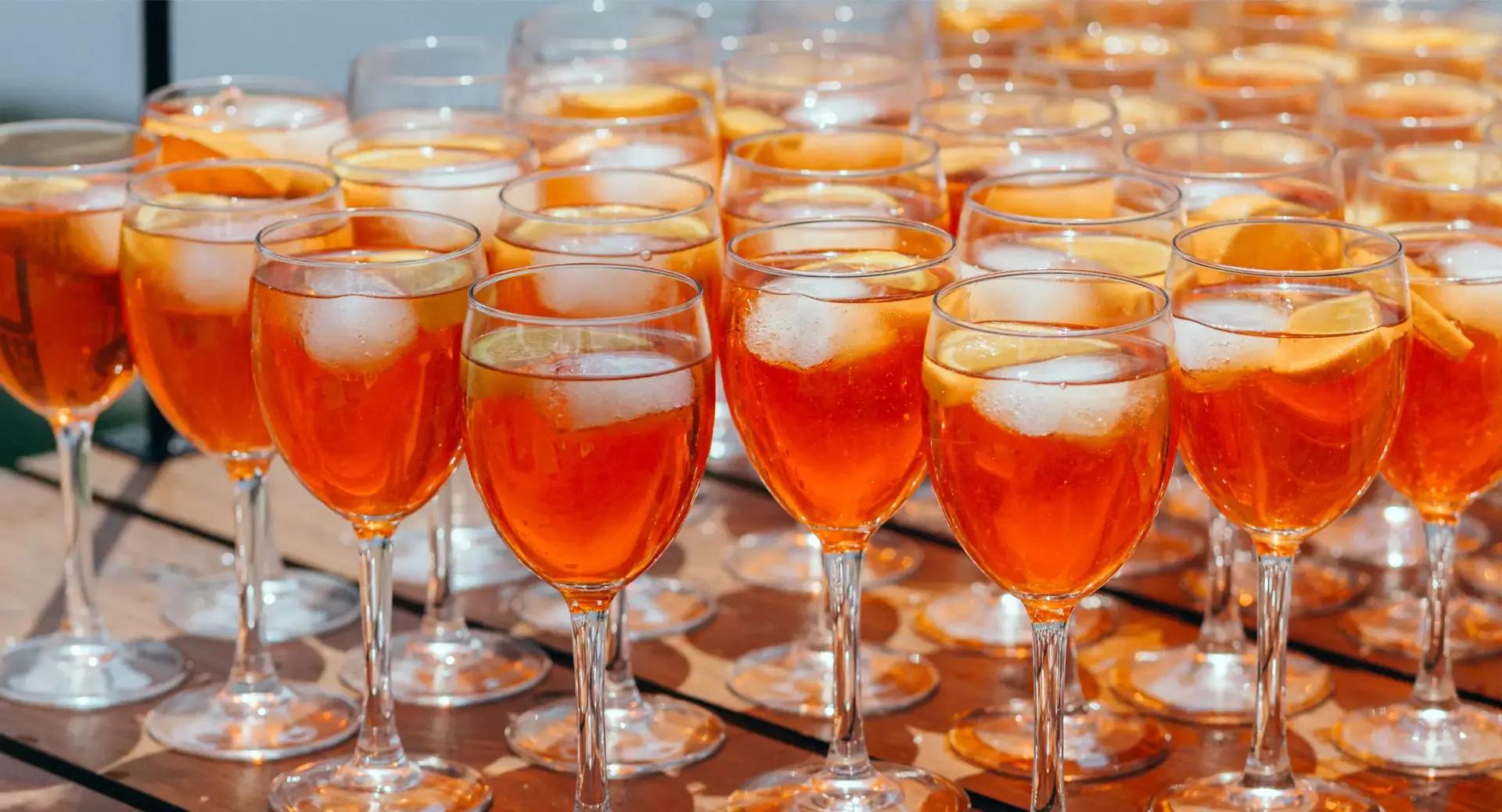 Multiple wine glasses filled with orange-colored drinks, ice cubes, and orange slices are arranged closely together on a wooden surface.