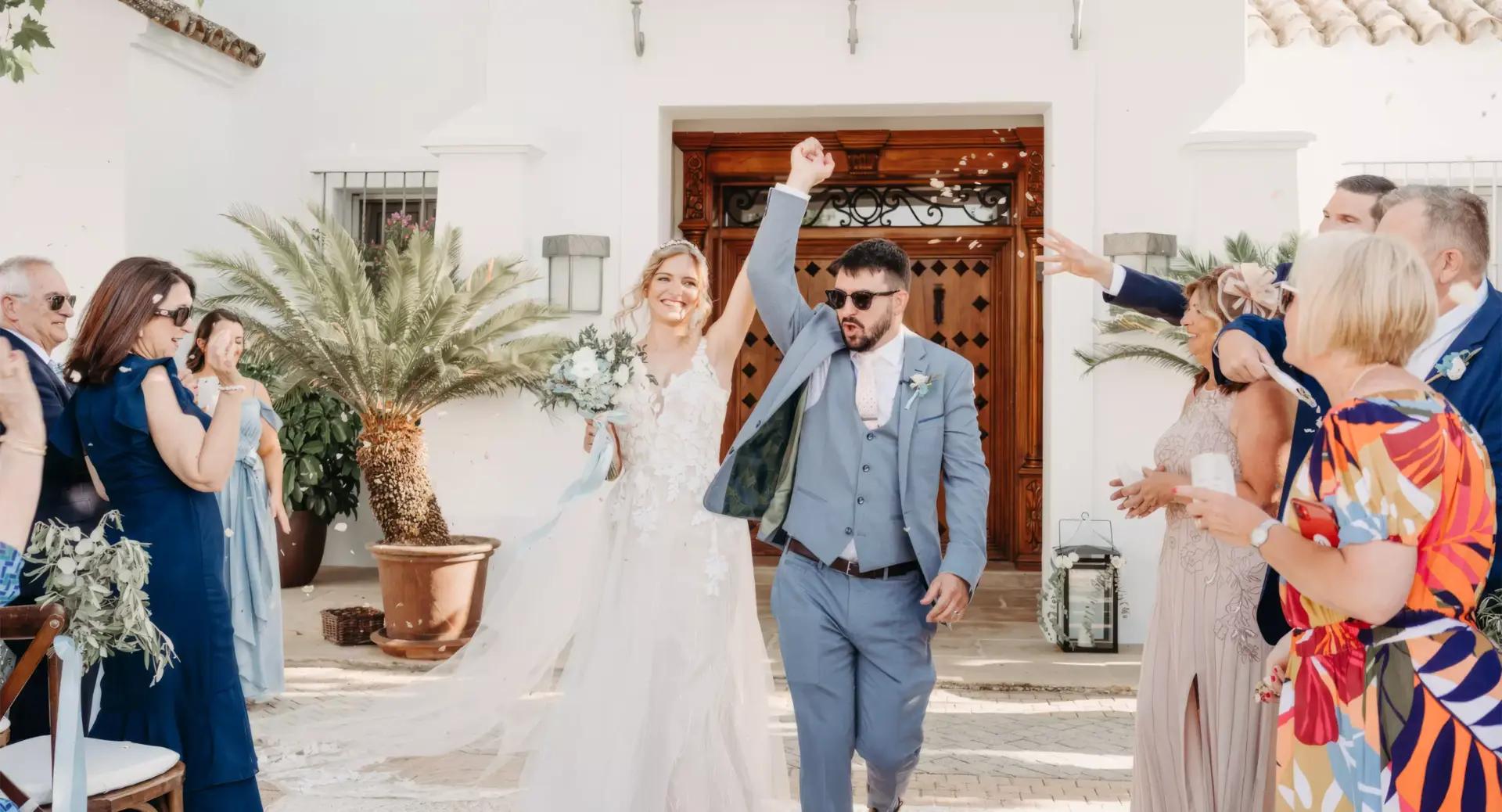 A bride and groom walk down an outdoor aisle as guests celebrate; the groom raises his arm in the air and the bride smiles, holding a bouquet.