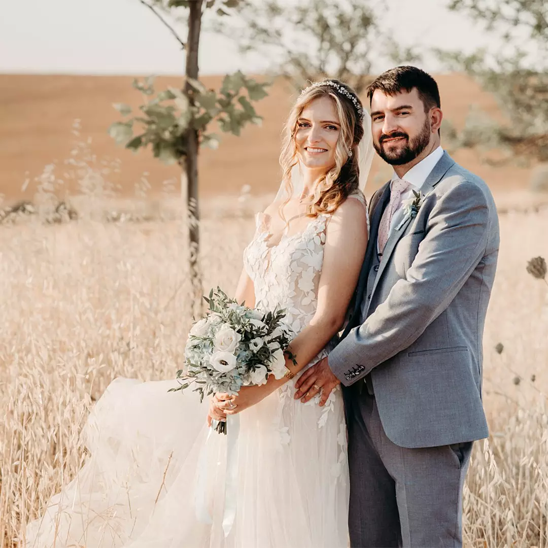 A bride in a white gown holding a bouquet stands beside a groom in a gray suit, posing together outdoors in a dry grassy field.